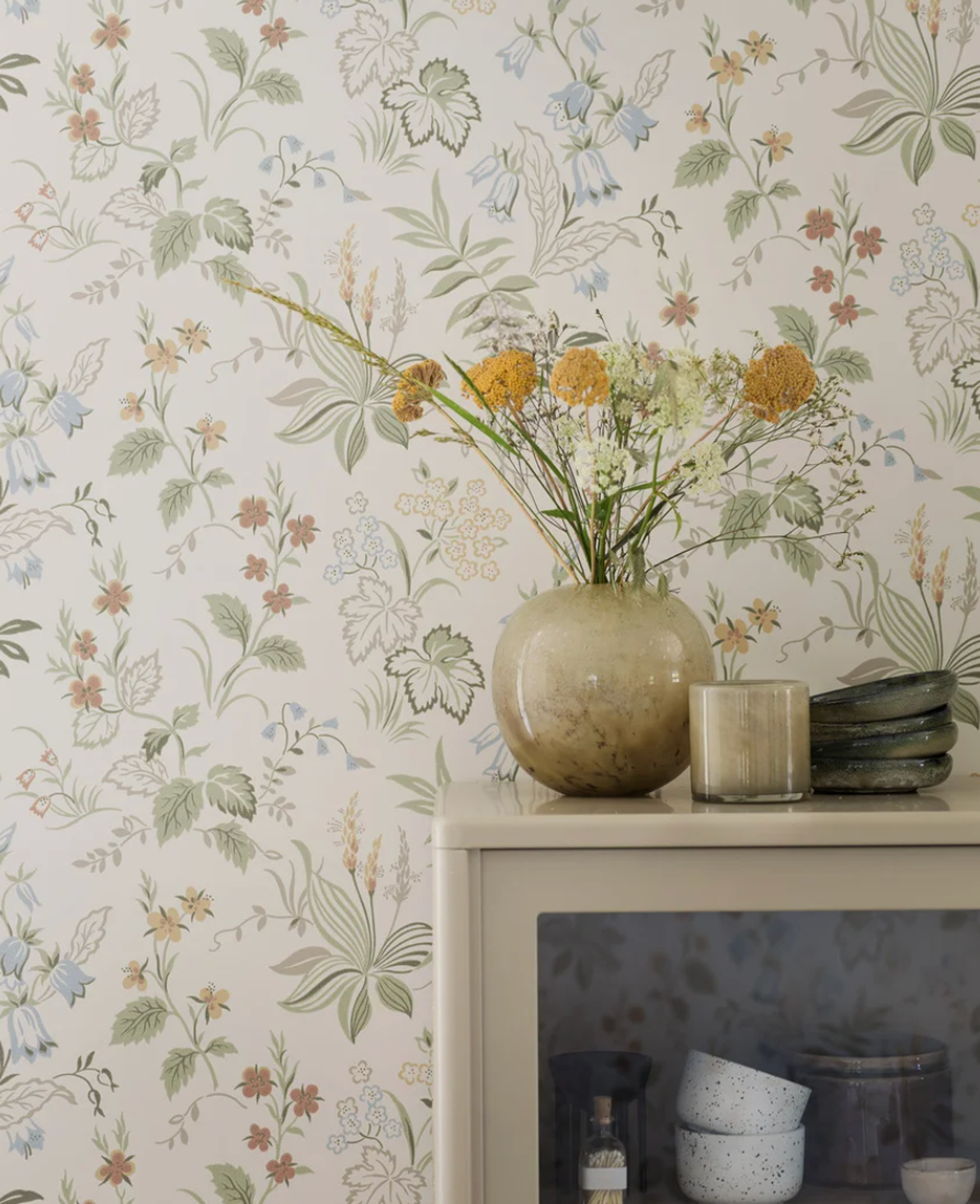 A light cabinet with a vase of flowers, a candle, and stacked plates, against a floral wallpaper.