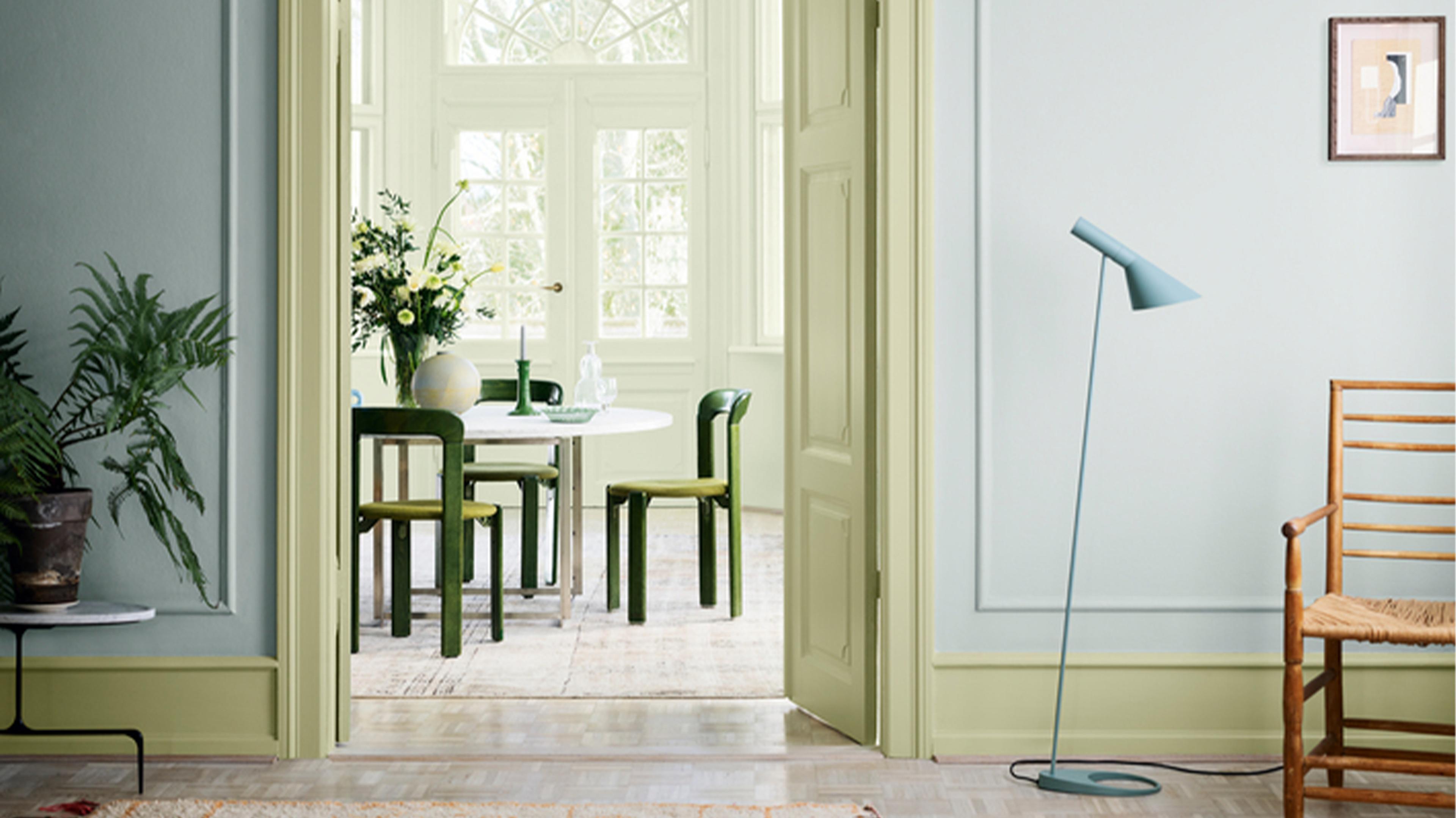 A bright interior showing two rooms connected by an open doorway. The foreground features pale blue and green walls with a blue floor lamp and wooden chair, while the background reveals a pale yellow dining room with green chairs.