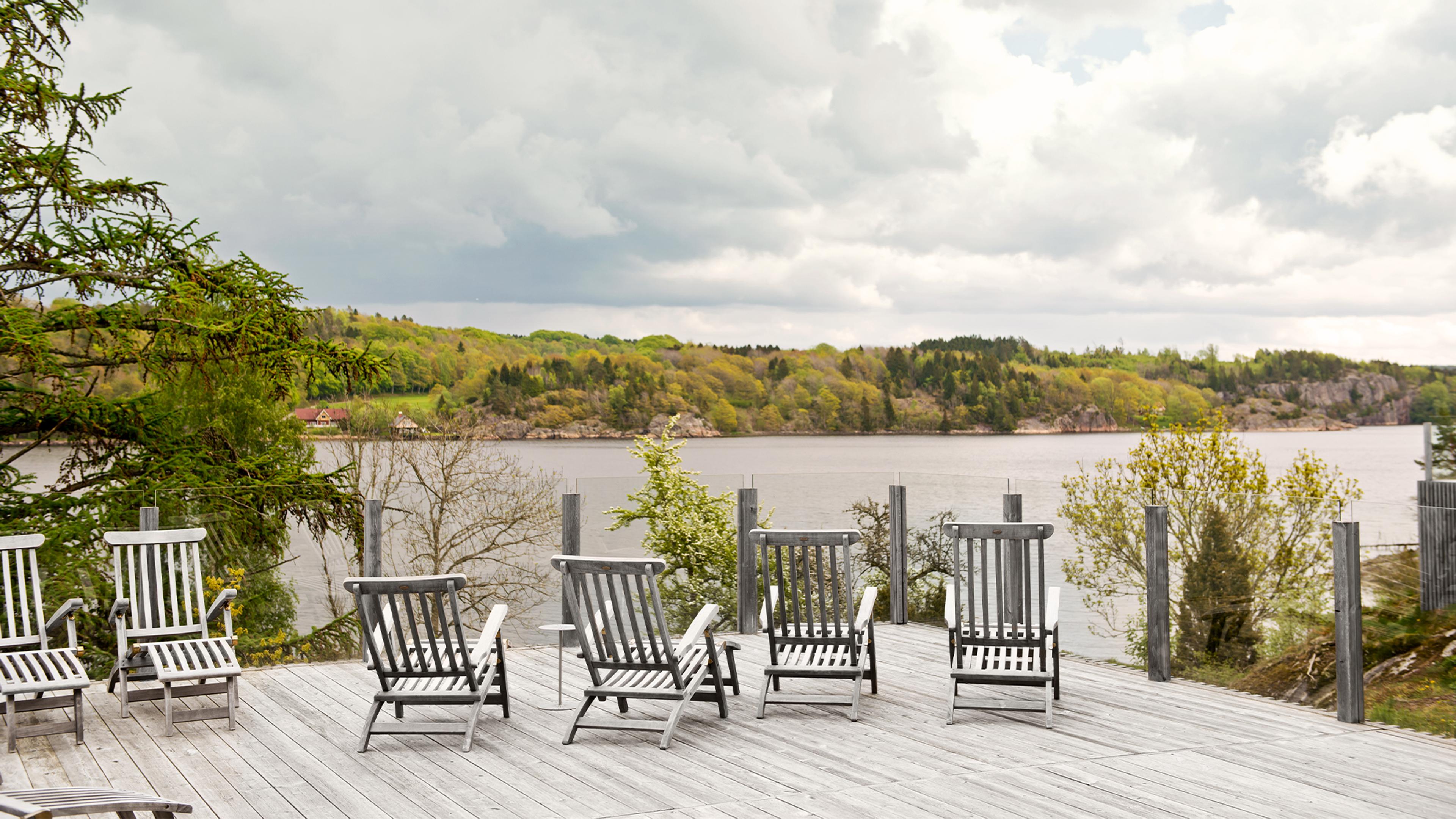 A wooden deck with empty lounge chairs overlooks a lake with forested hills under a cloudy sky.