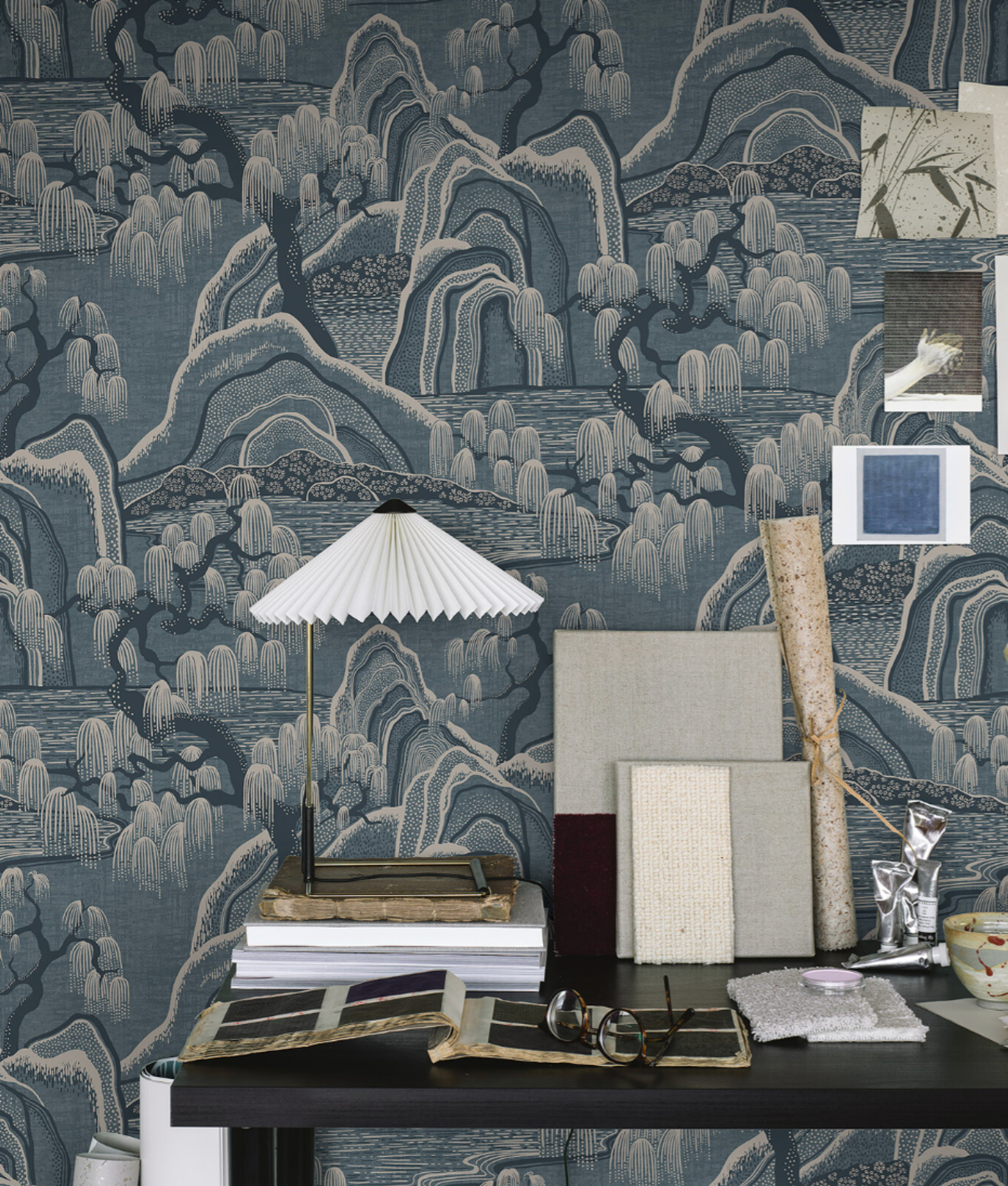 Blue East Asian landscape wallpaper with mountains and willows behind a desk holding a pleated lamp, books, and fabric swatches.