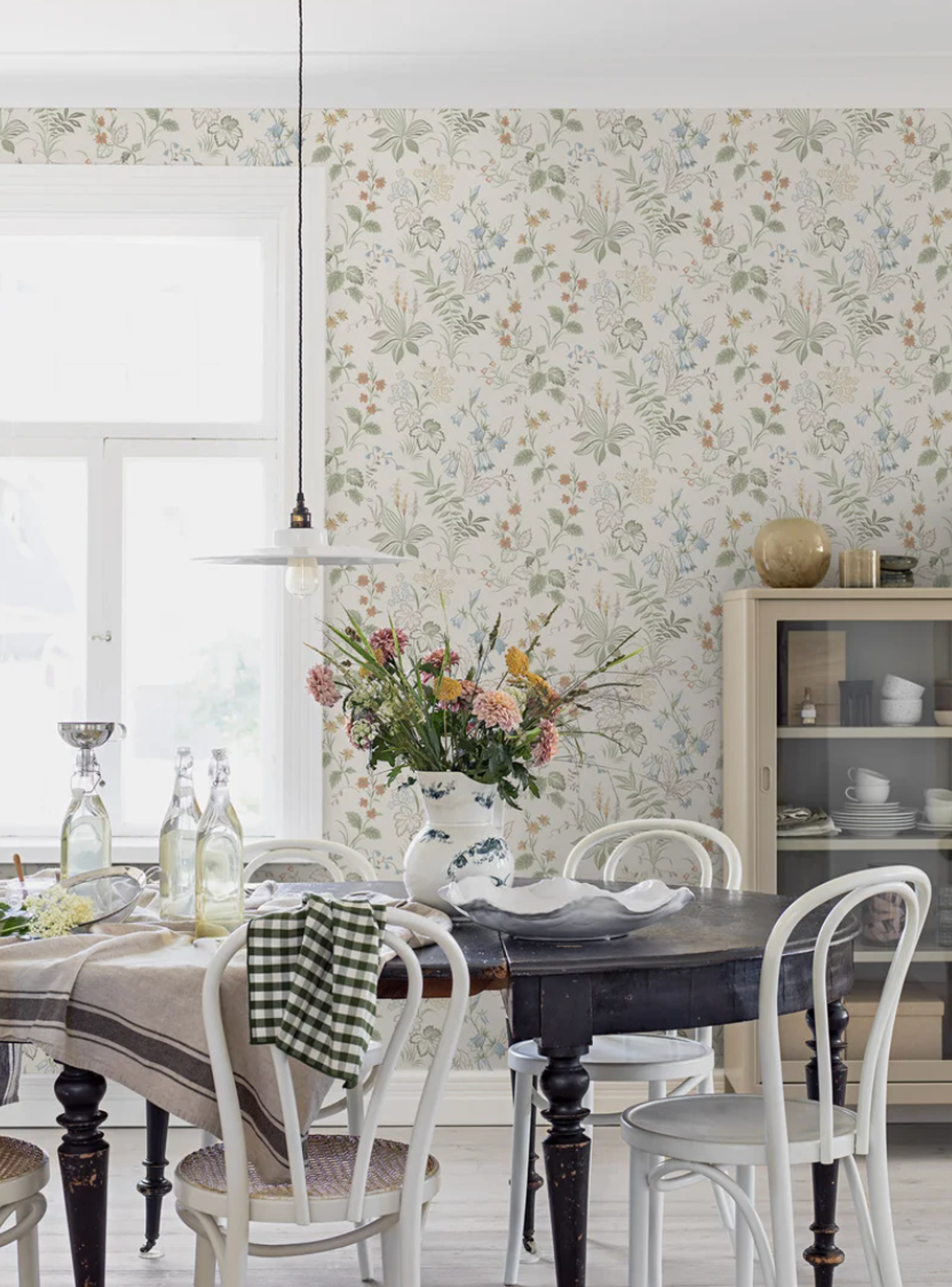 A dining room with light floral wallpaper, a dark wood dining table set with a vase of flowers, and white chairs.