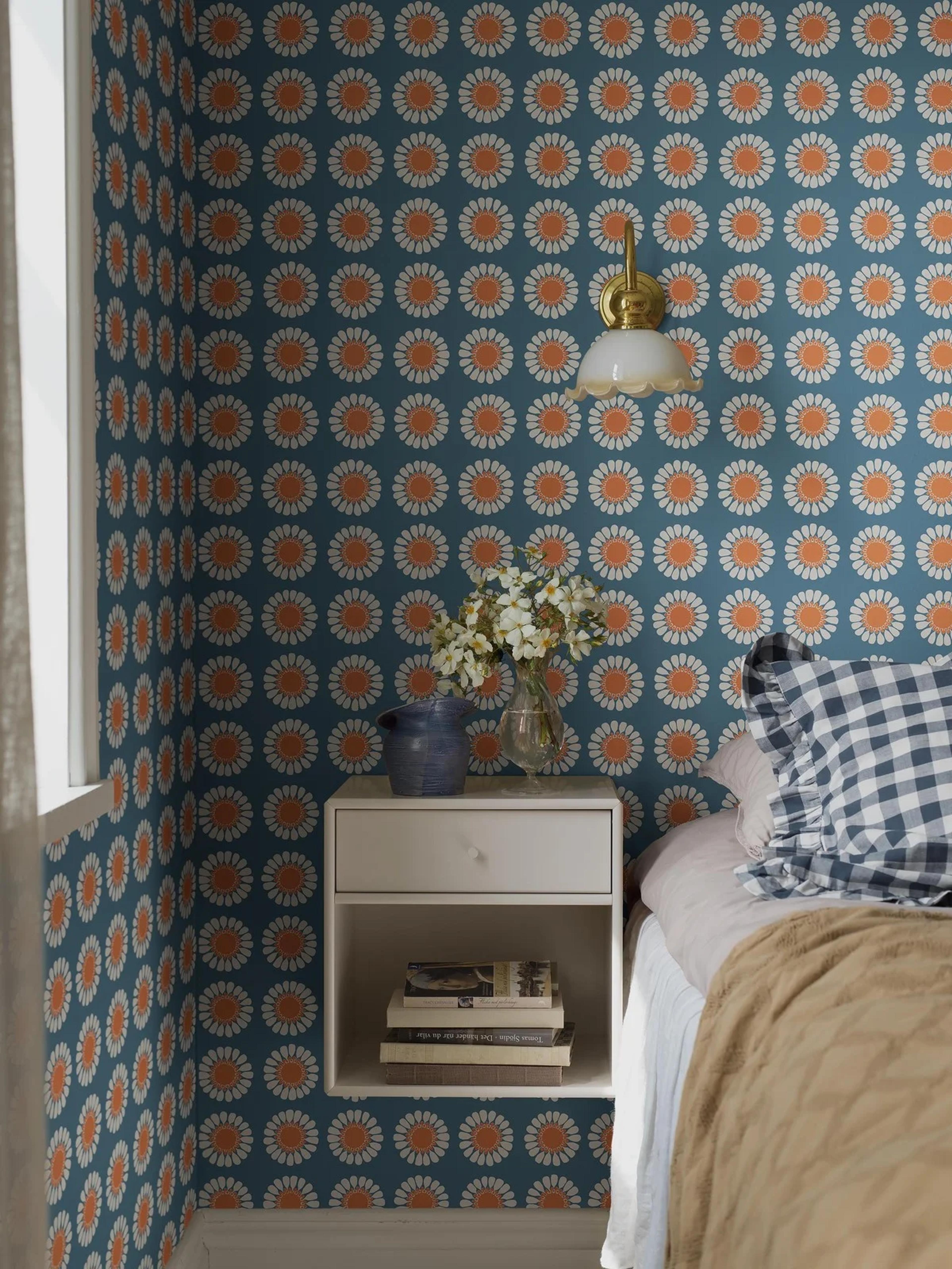 A bedroom with teal wallpaper featuring white and orange daisy patterns. A brass sconce hangs above a white floating nightstand beside a bed with a gingham pillow.