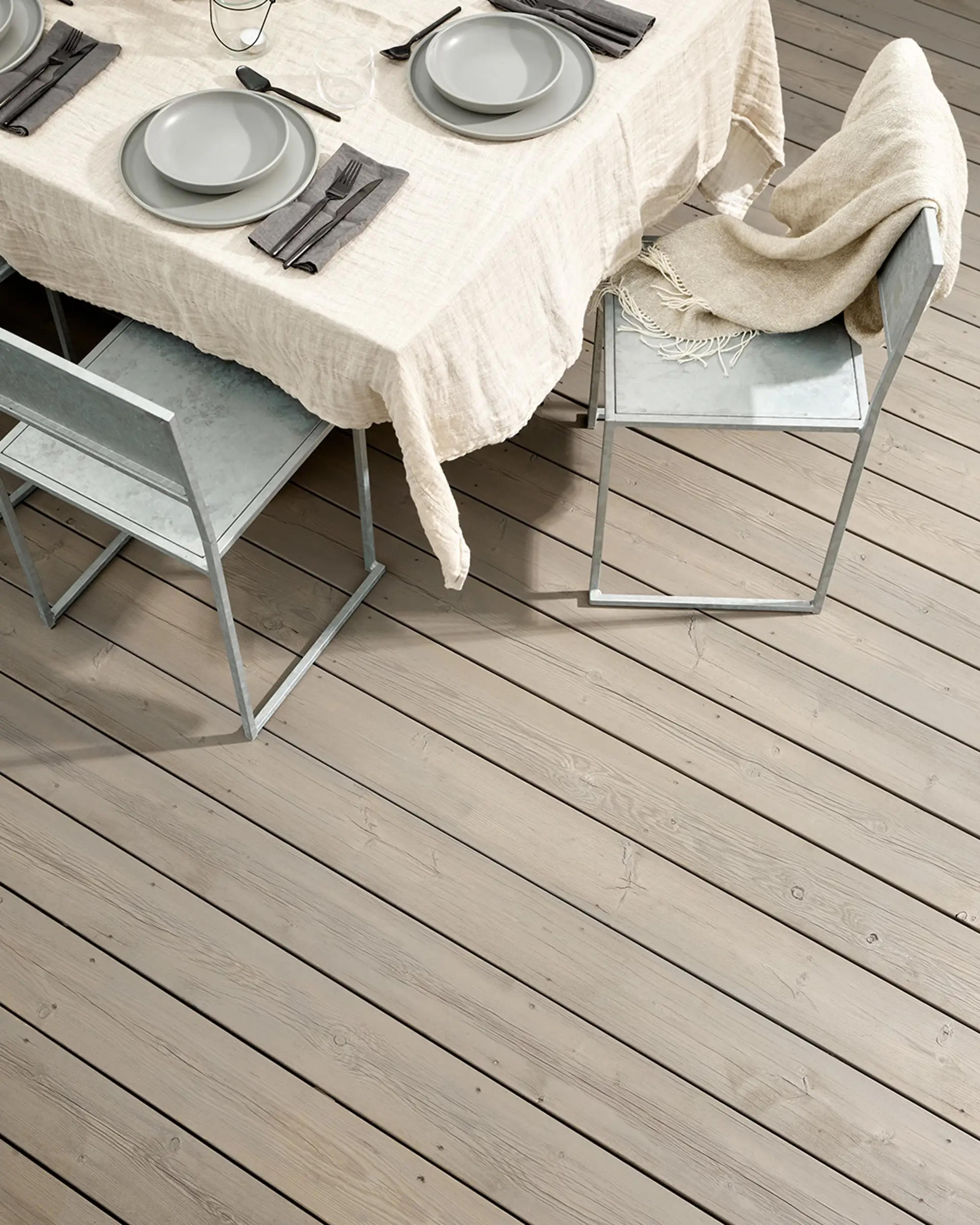 A dining table with grey place settings on a light tablecloth, flanked by grey metal chairs on a light wooden deck. One chair has a beige blanket.