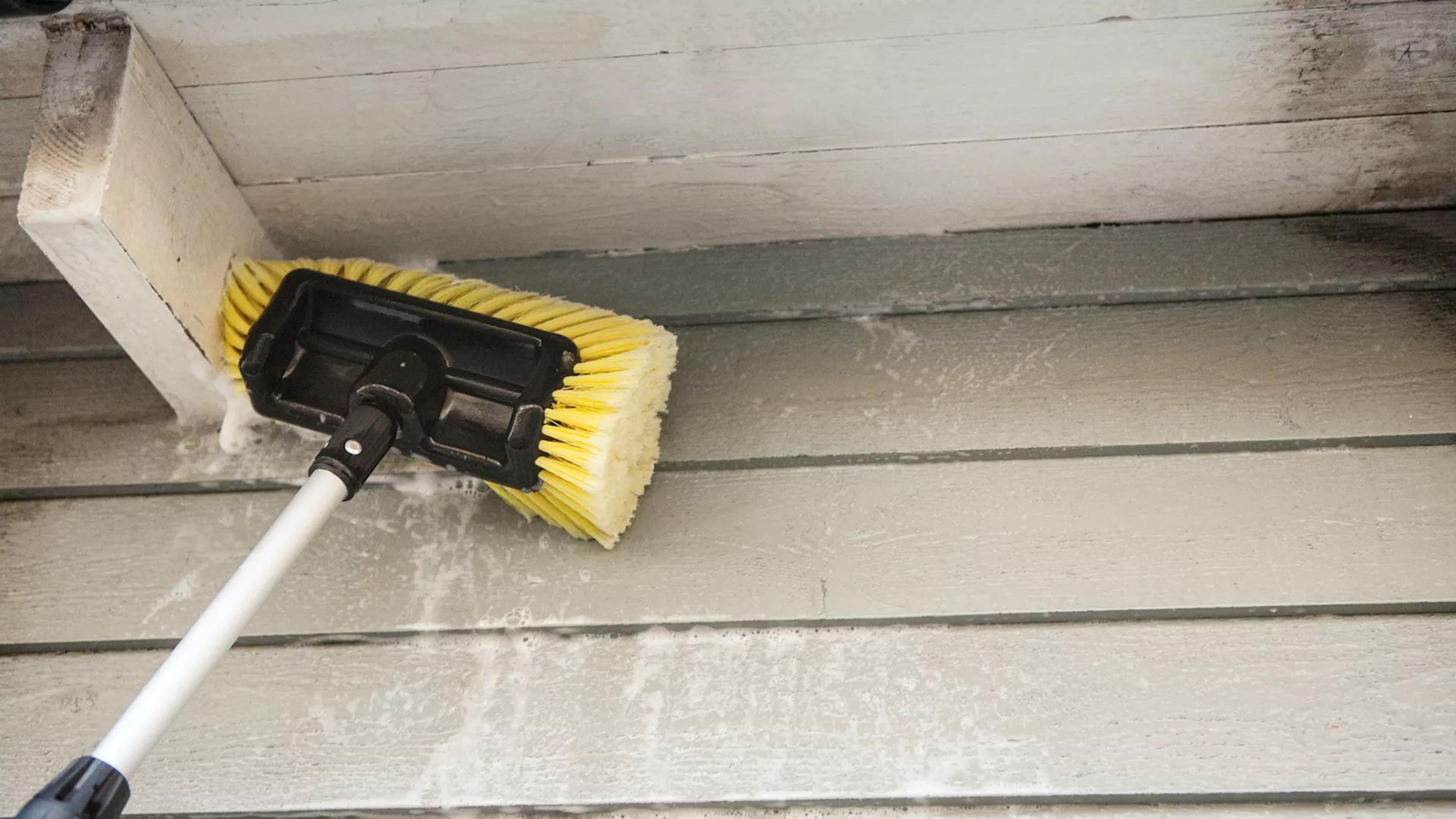 A long-handled yellow brush cleaning light green house siding with soap.