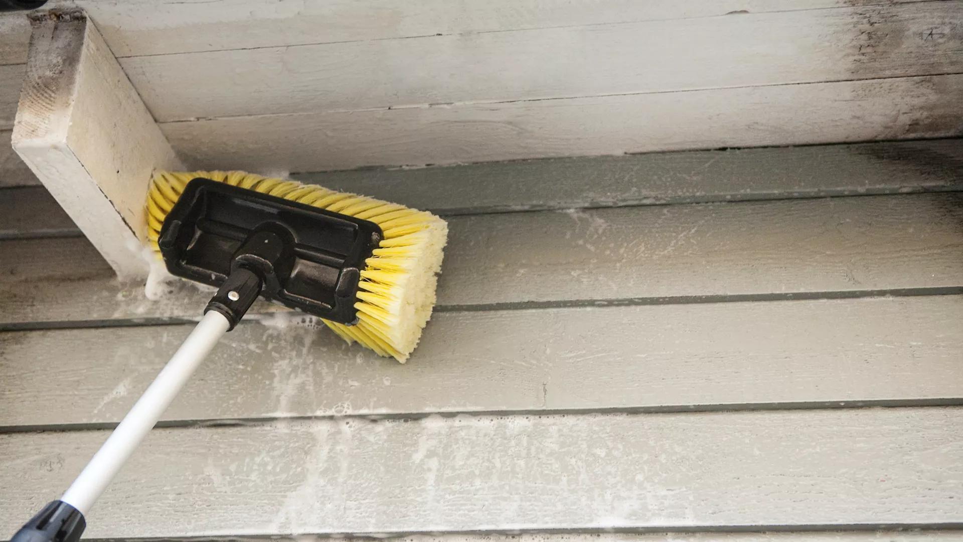 A long-handled yellow brush cleaning light green house siding with soap.
