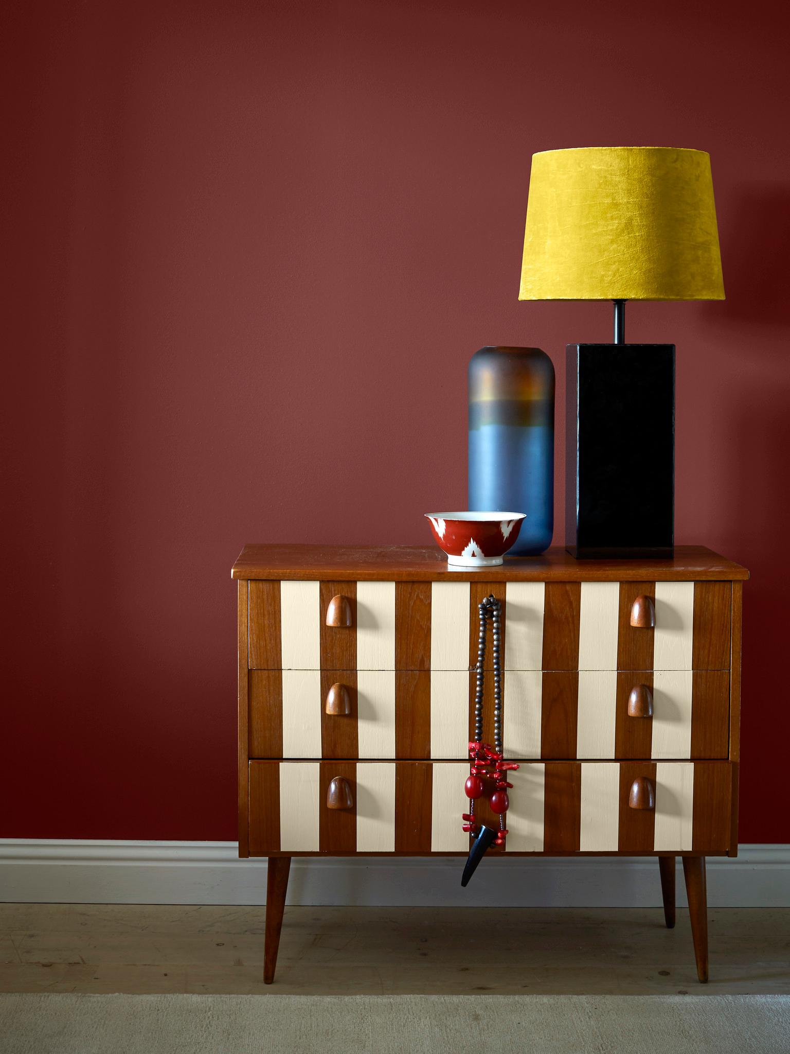 A striped wooden dresser against a deep red wall, topped with a yellow lamp, a blue vase, and a small bowl, with a beaded necklace hanging from a drawer.