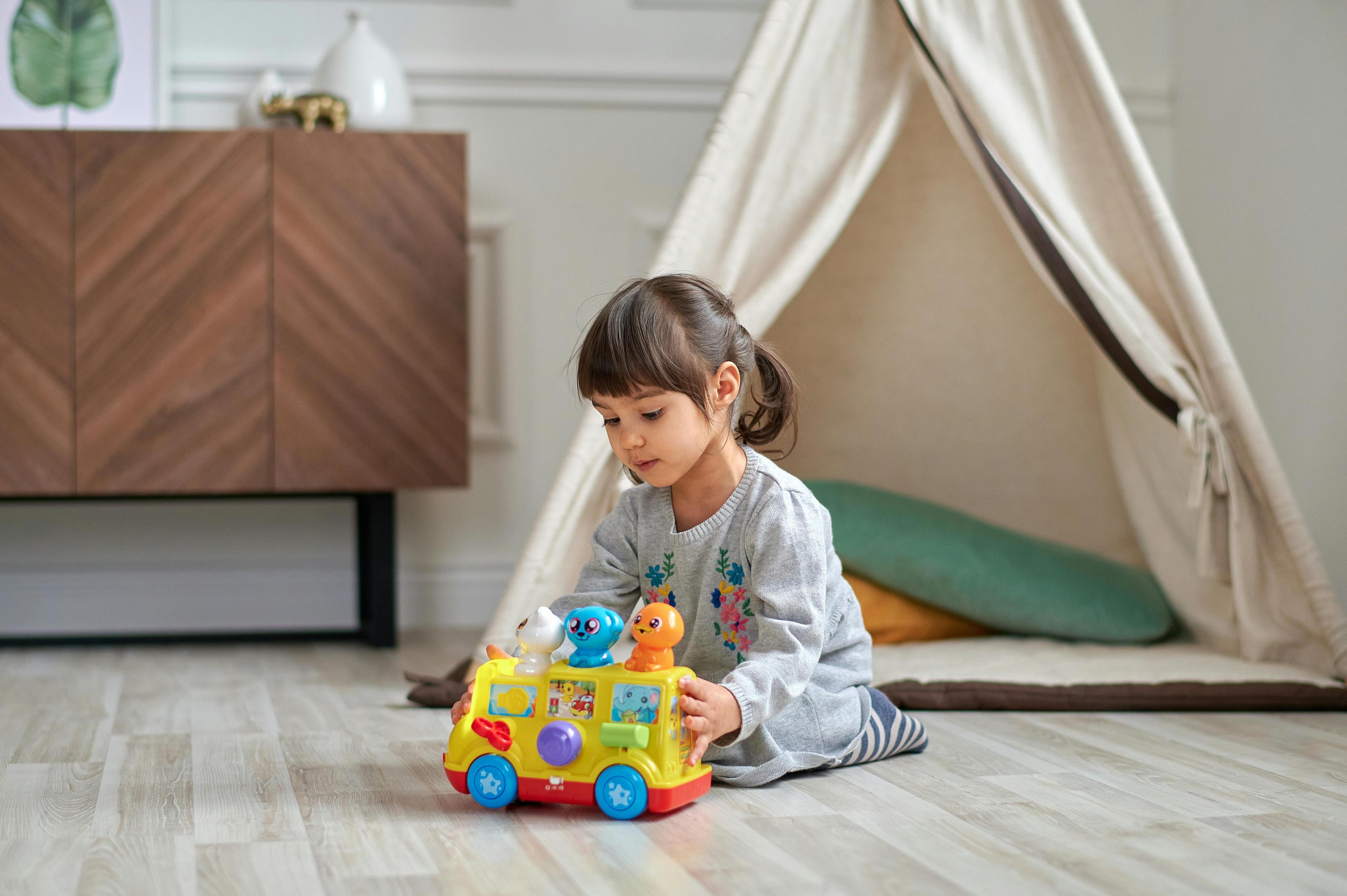 A young child plays with a yellow toy bus with animal figures on a light wooden floor.