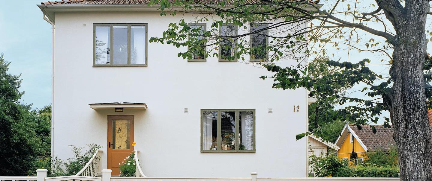 A white two-story house with green window frames, a wooden front door, and the number 12, behind a white fence and a tree.