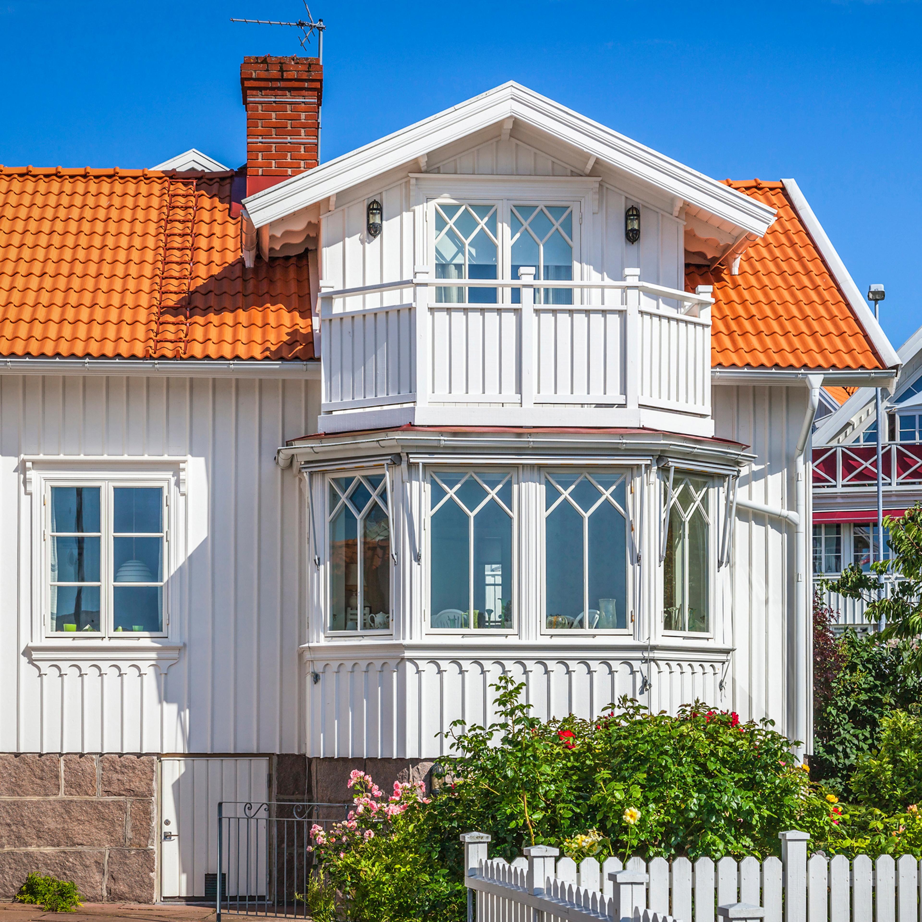A white wooden house with an orange tiled roof, a bay window, a balcony, green bushes, and a white picket fence.