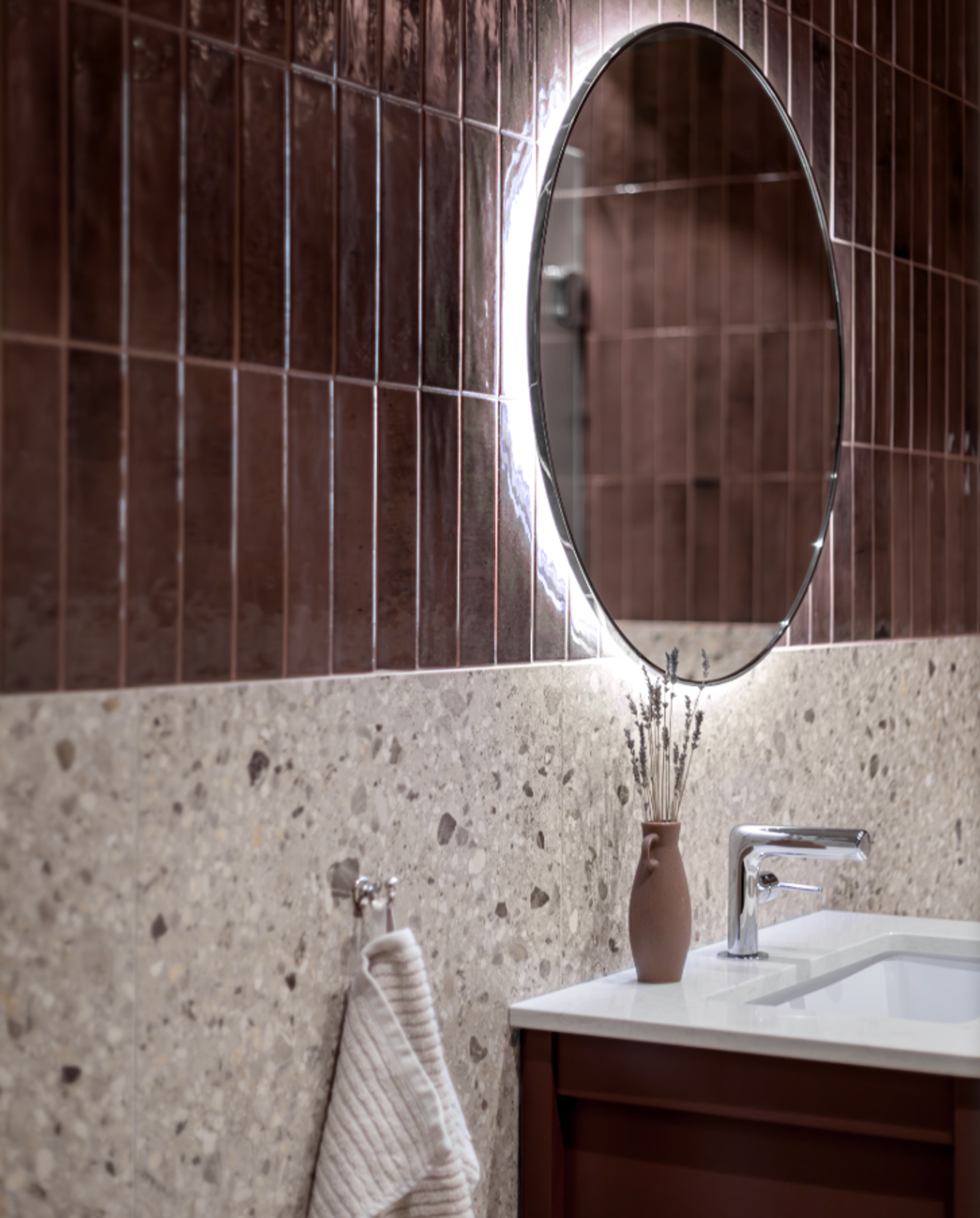 Modern bathroom with an oval backlit mirror, brown glossy subway tiles, grey terrazzo tiles, and a white sink.