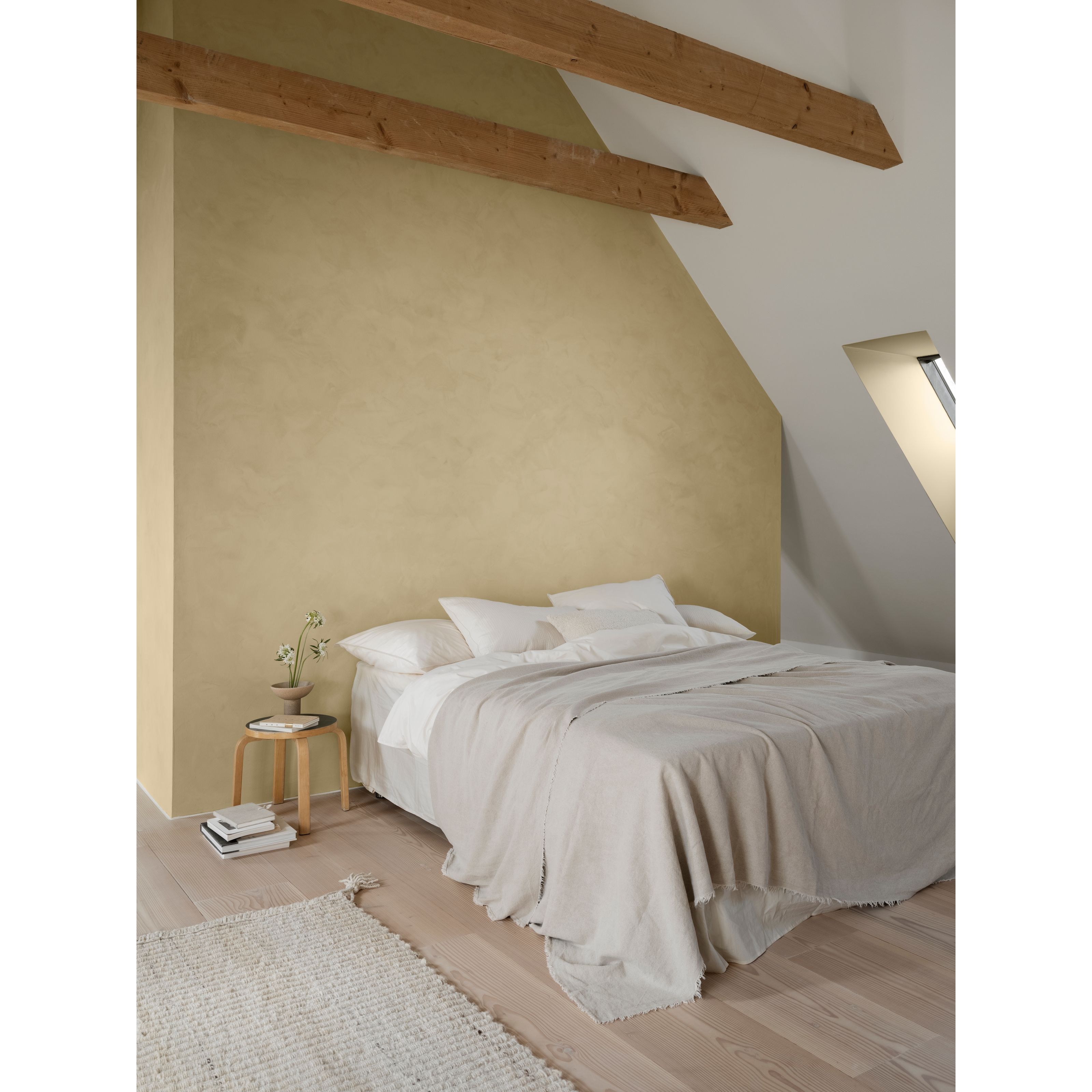 A minimalist bedroom with light yellow textured walls, exposed wooden beams, a bed with white bedding, and a skylight.