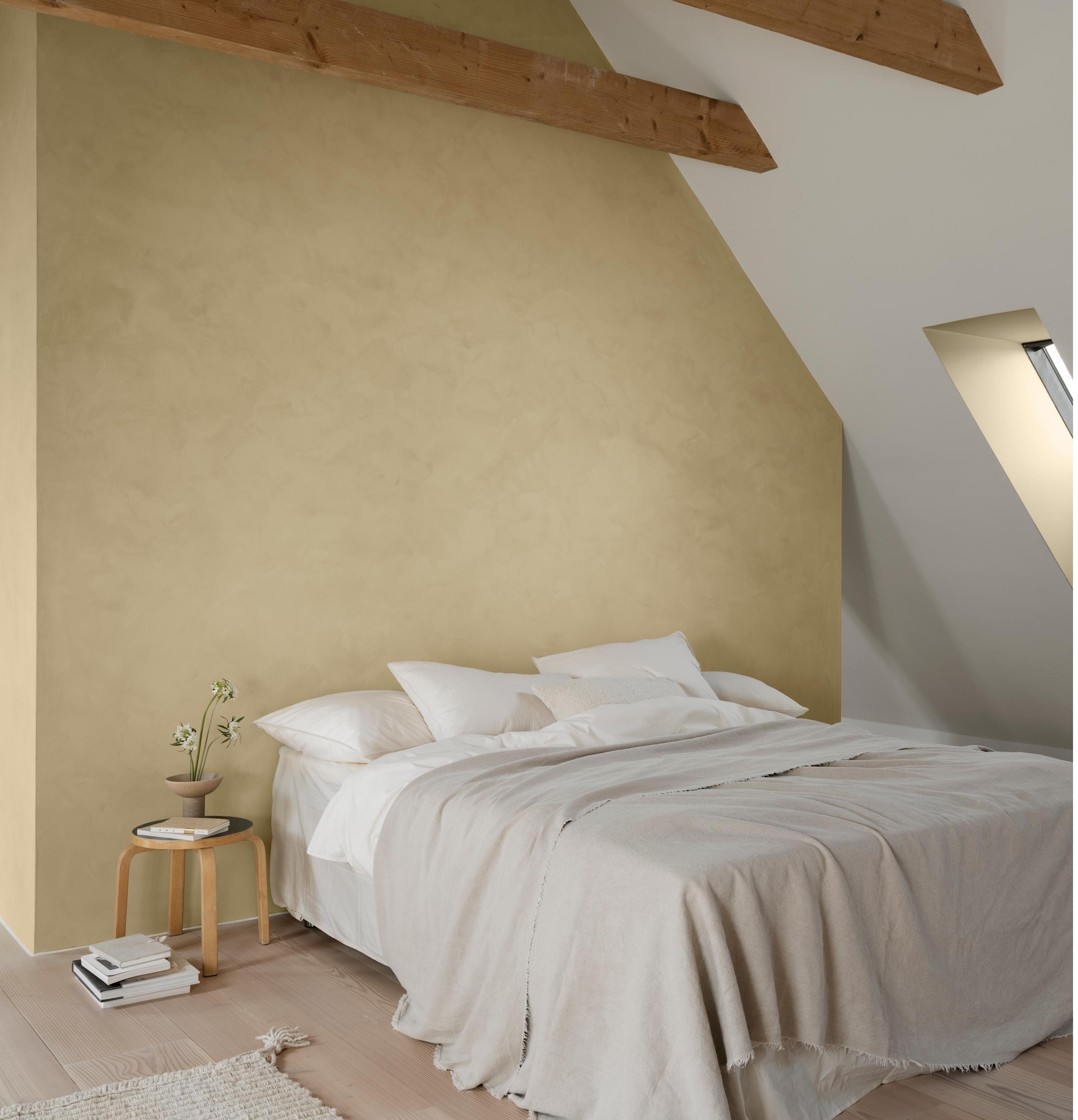 A minimalist bedroom with light yellow textured walls, exposed wooden beams, a bed with white bedding, and a skylight.