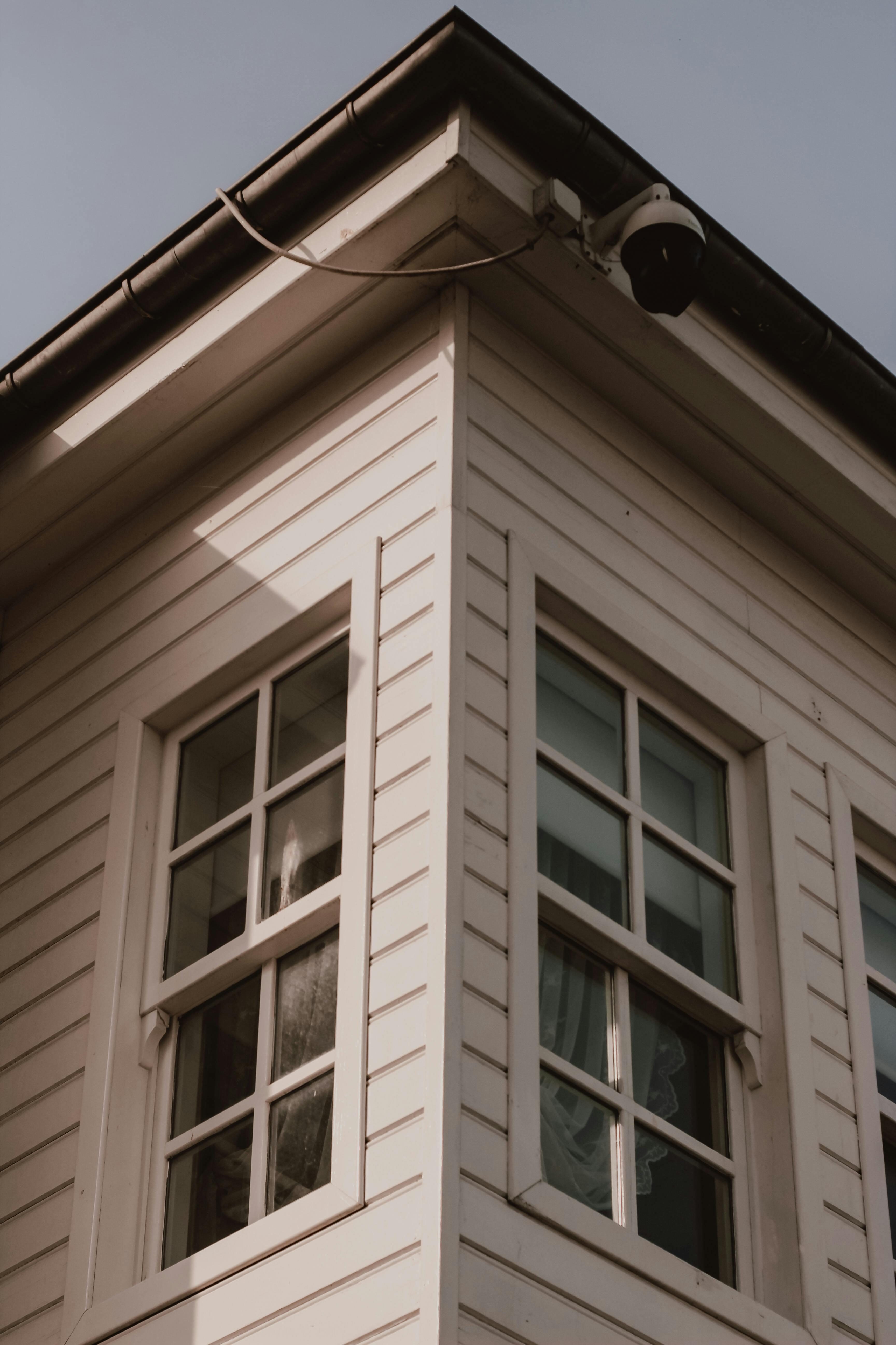 Corner of a light-colored building with horizontal siding, two windows, and a security camera.