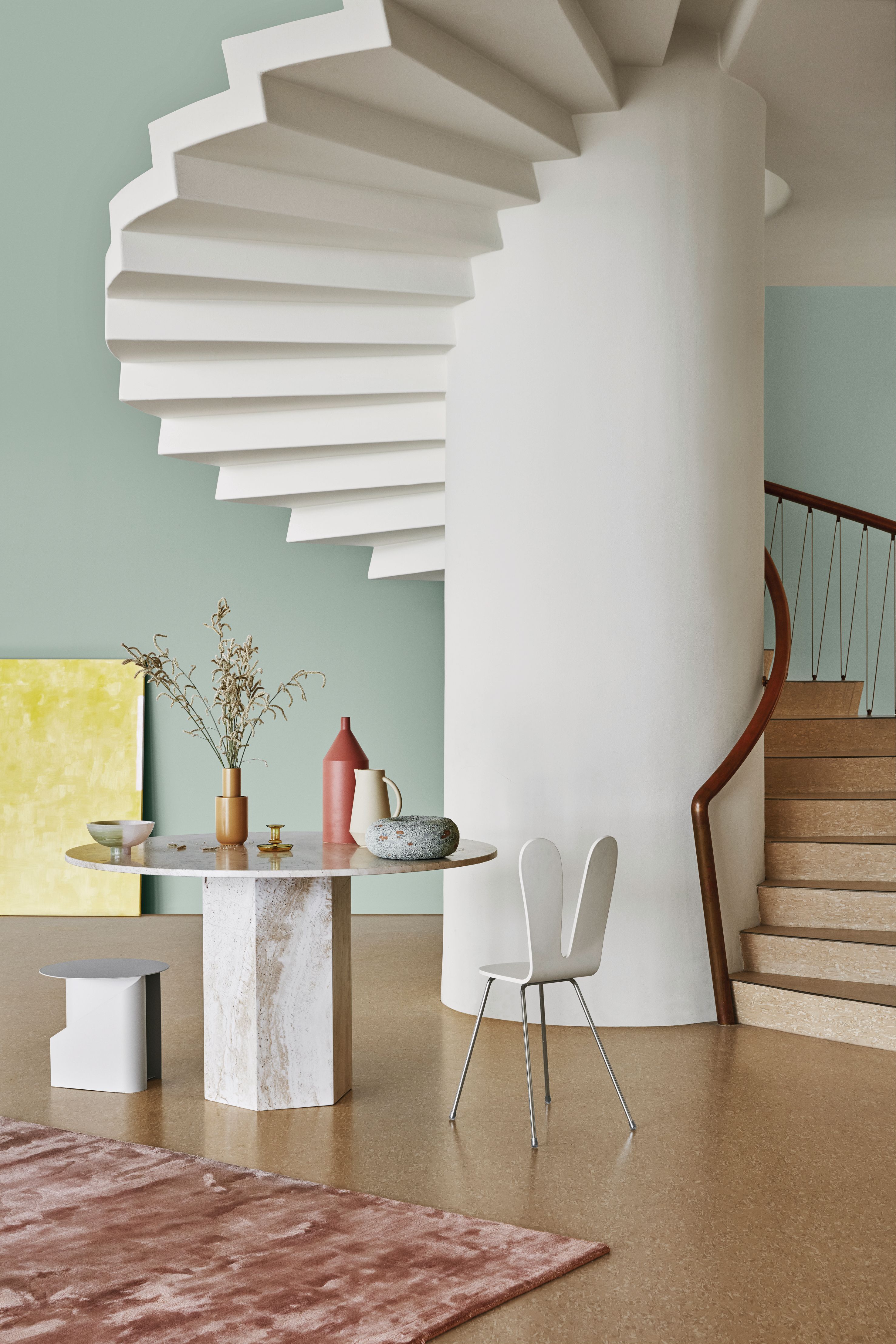 A modern interior with a white spiral staircase, a round marble table with decor, and a wooden staircase, all against light green walls and a speckled floor.