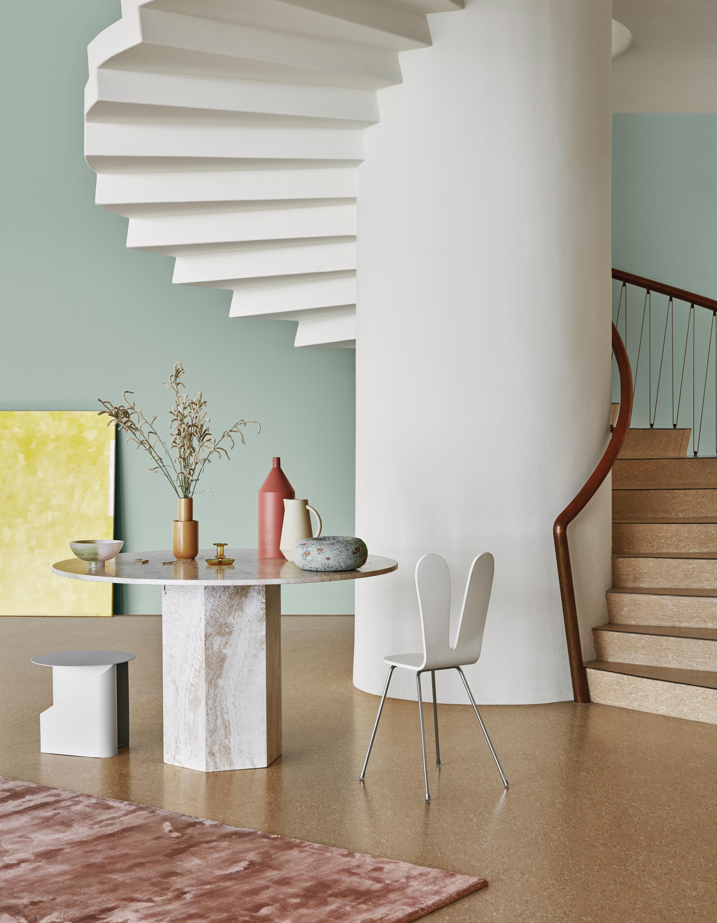 A modern interior with a white spiral staircase, a round marble table with decor, and a wooden staircase, all against light green walls and a speckled floor.