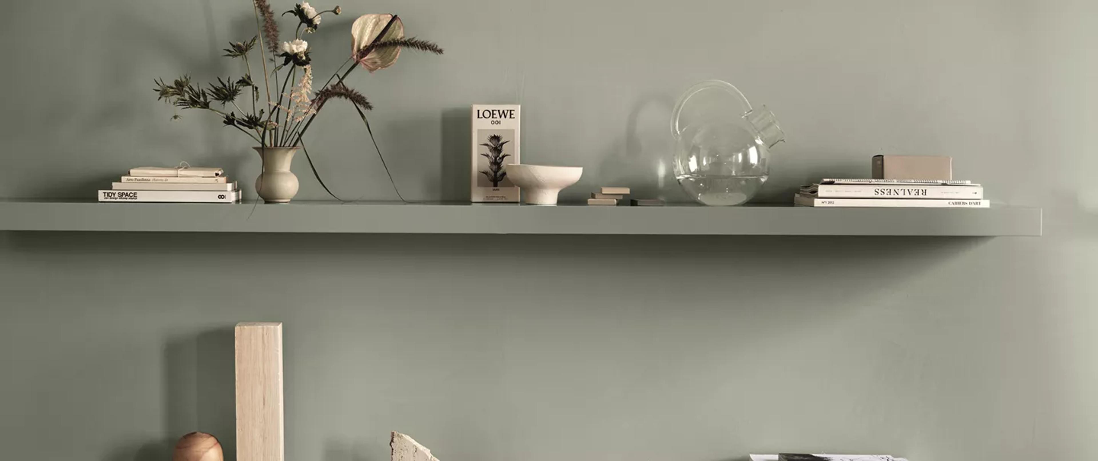 A minimalist floating shelf on a sage green wall, styled with books, dried flowers in a vase, and a glass carafe.