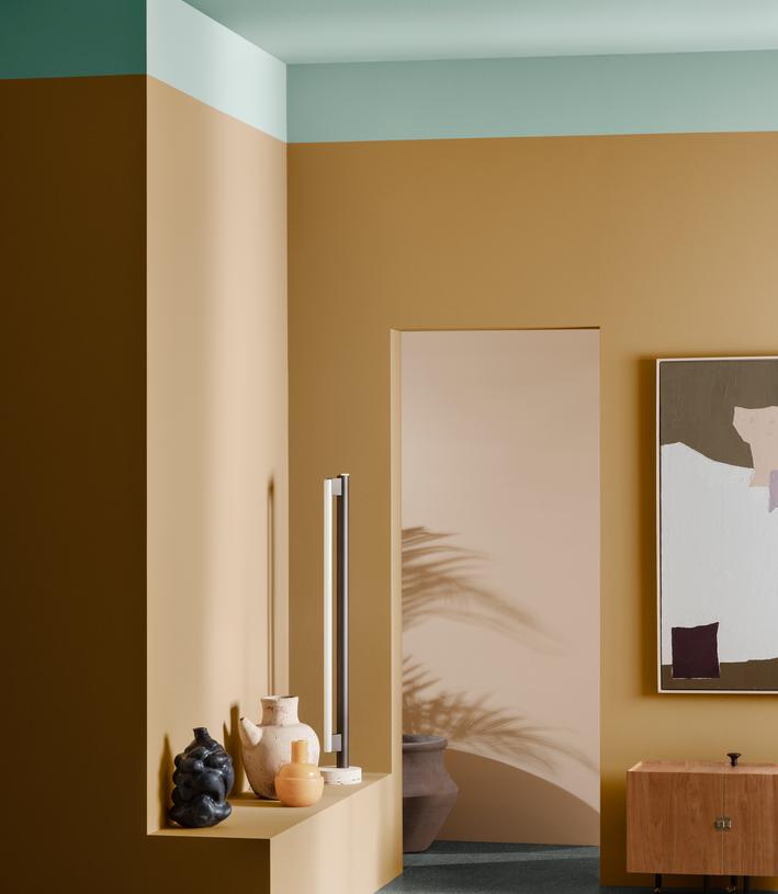 A stylish room with warm ochre walls, a mint green ceiling, and dark grey floors. A modern lamp and decorative vases sit on a built-in ledge, while an abstract painting and wooden cabinet adorn the right wall.