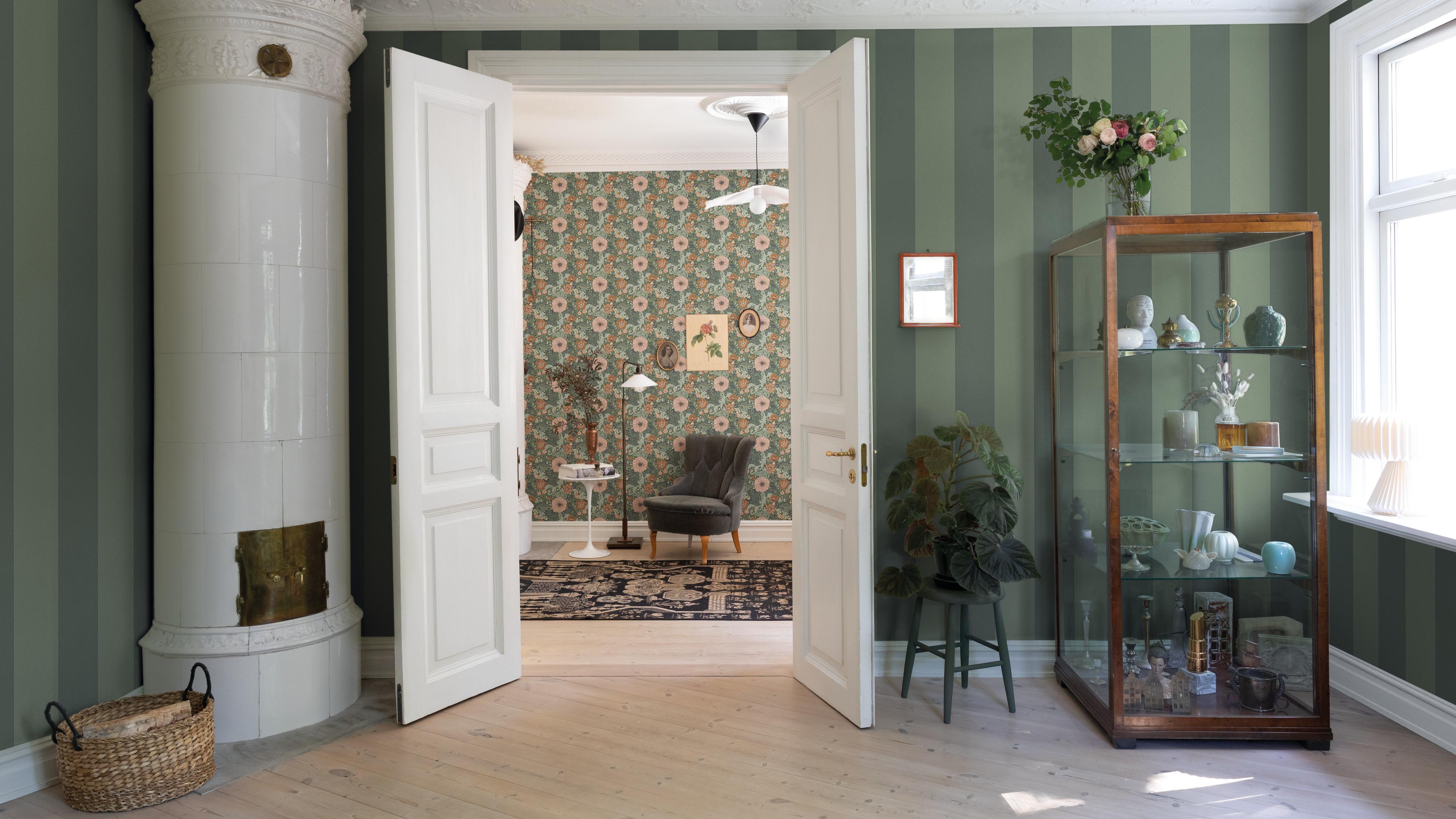 A green striped room with a white tiled stove and a display cabinet, looking through an open door into a floral-patterned room.