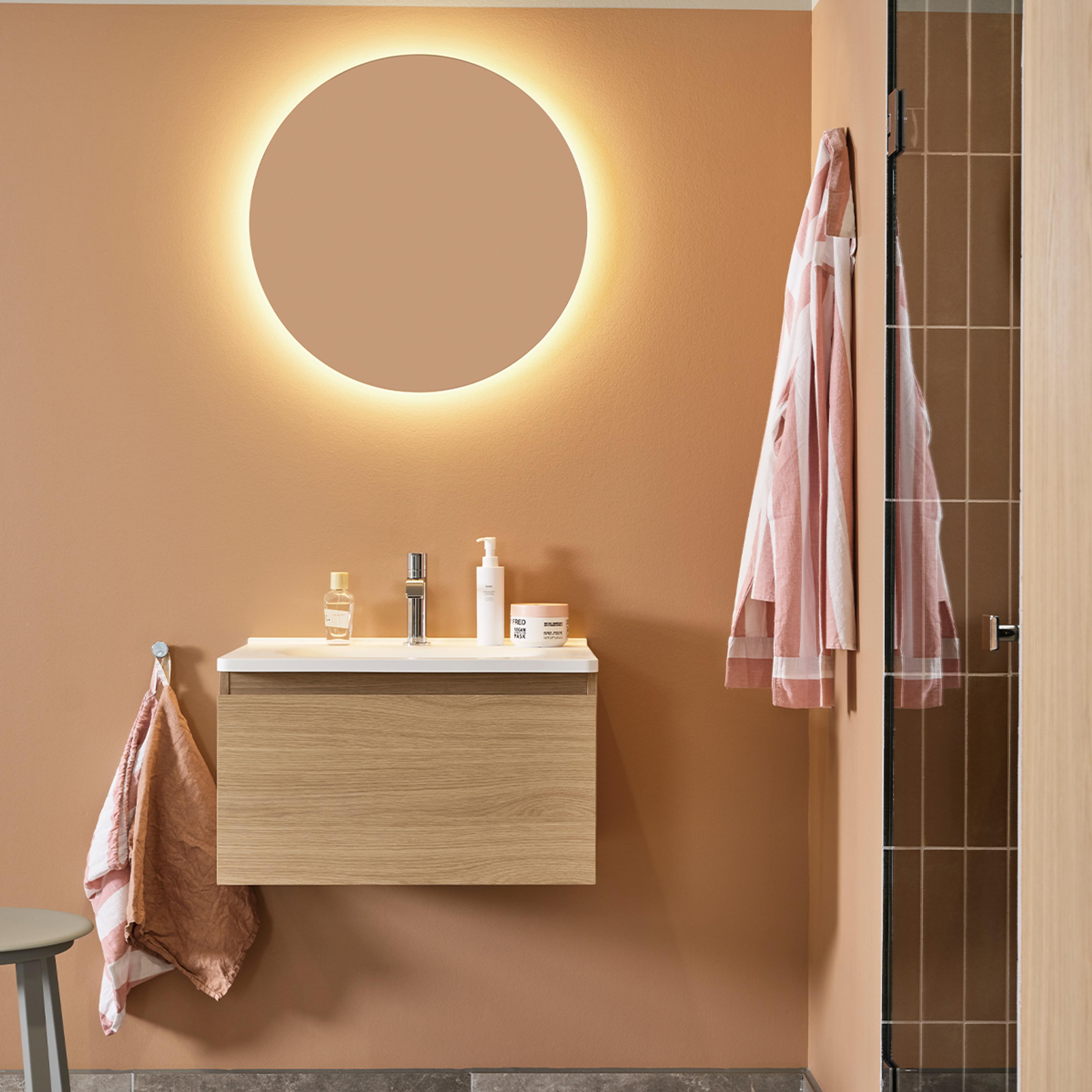 A modern bathroom with a round backlit mirror above a wooden floating vanity with a white sink, against terracotta walls, and hanging towels.