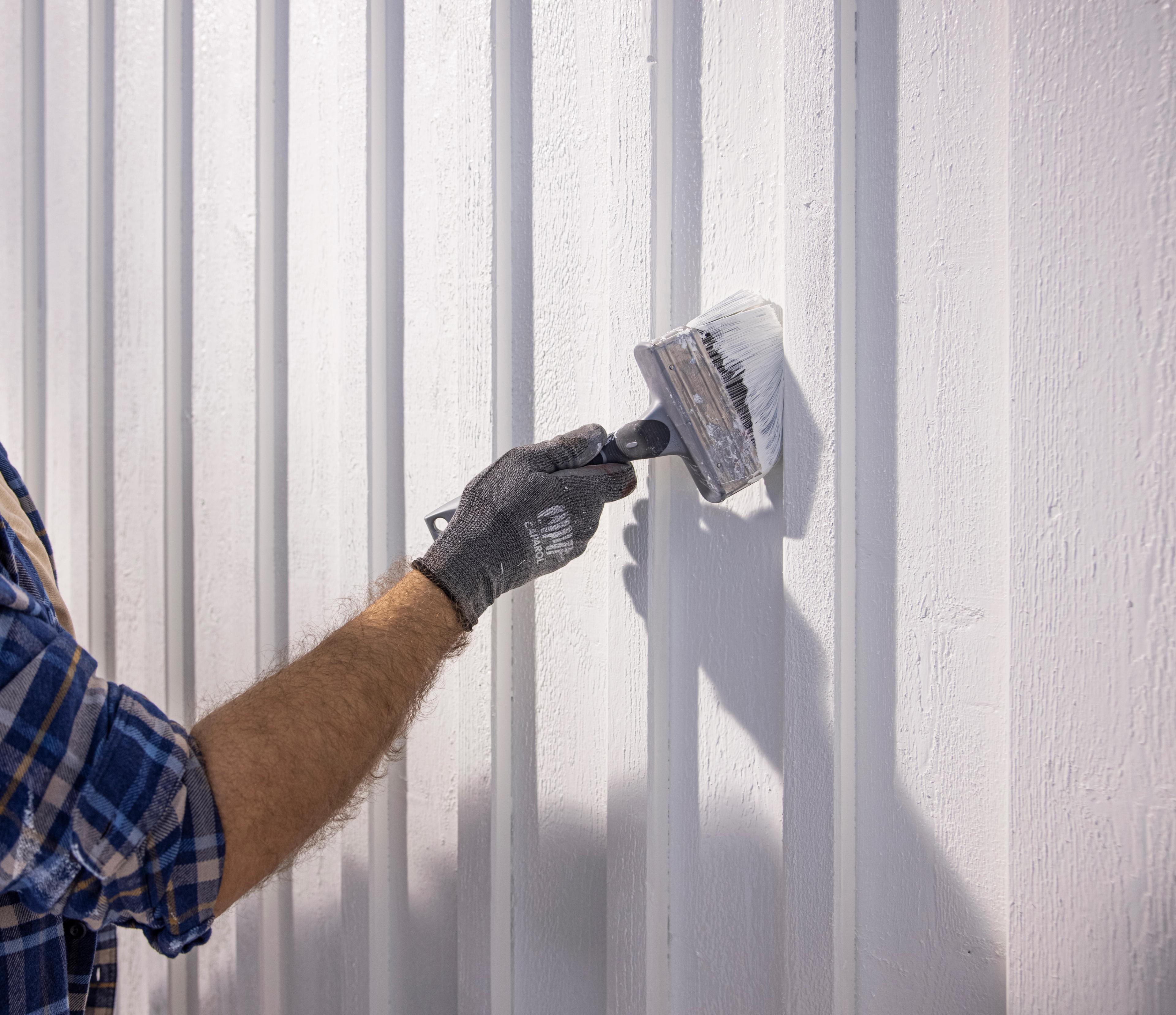Gloved hand painting a white paneled wall with a wide brush.