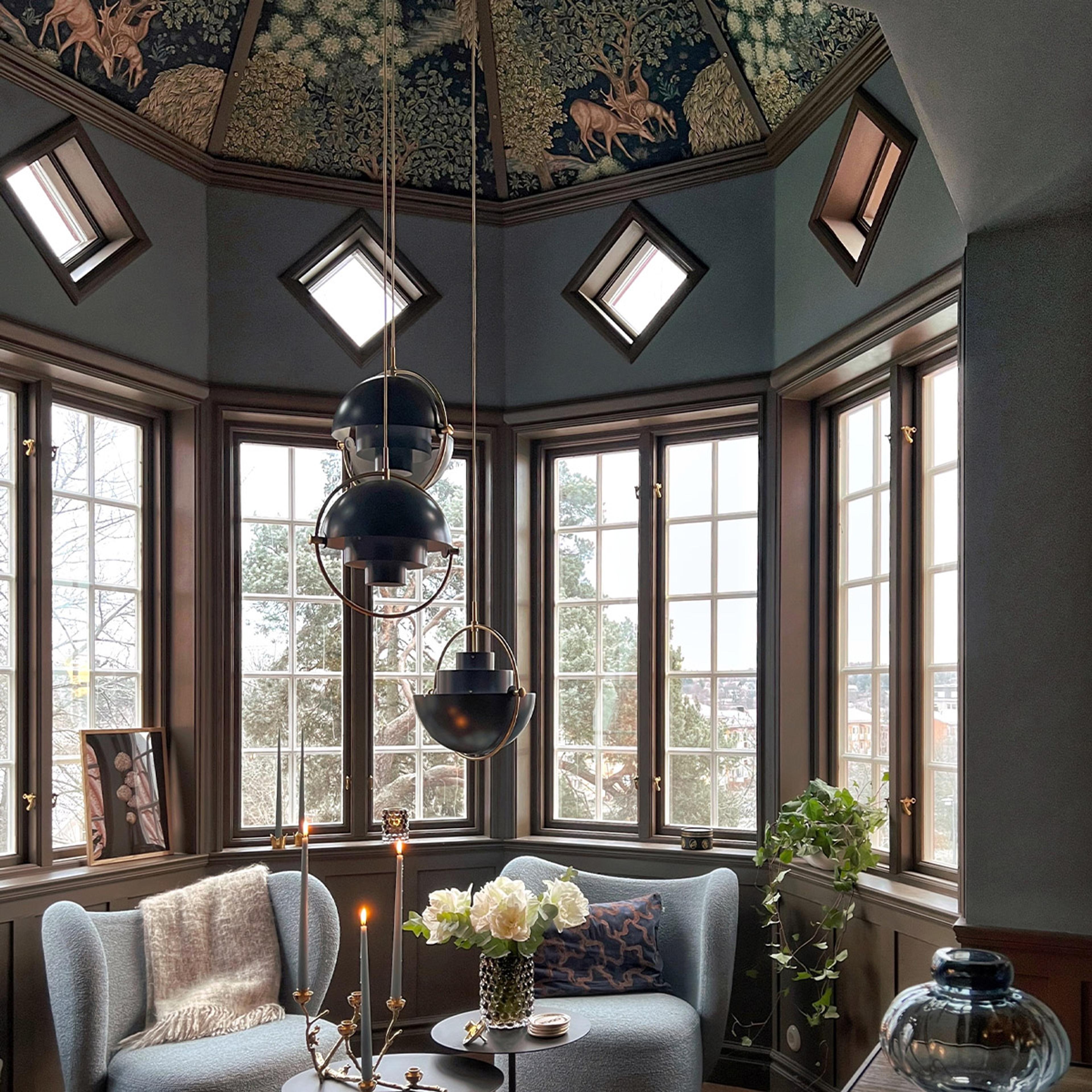 A luxurious room with a domed ceiling featuring a dark forest and deer tapestry, large bay windows, three black geometric pendant lights, and two light blue armchairs around a small table with flowers and candles.