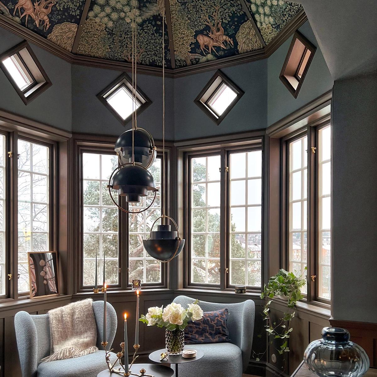 A luxurious room with a domed ceiling featuring a dark forest and deer tapestry, large bay windows, three black geometric pendant lights, and two light blue armchairs around a small table with flowers and candles.