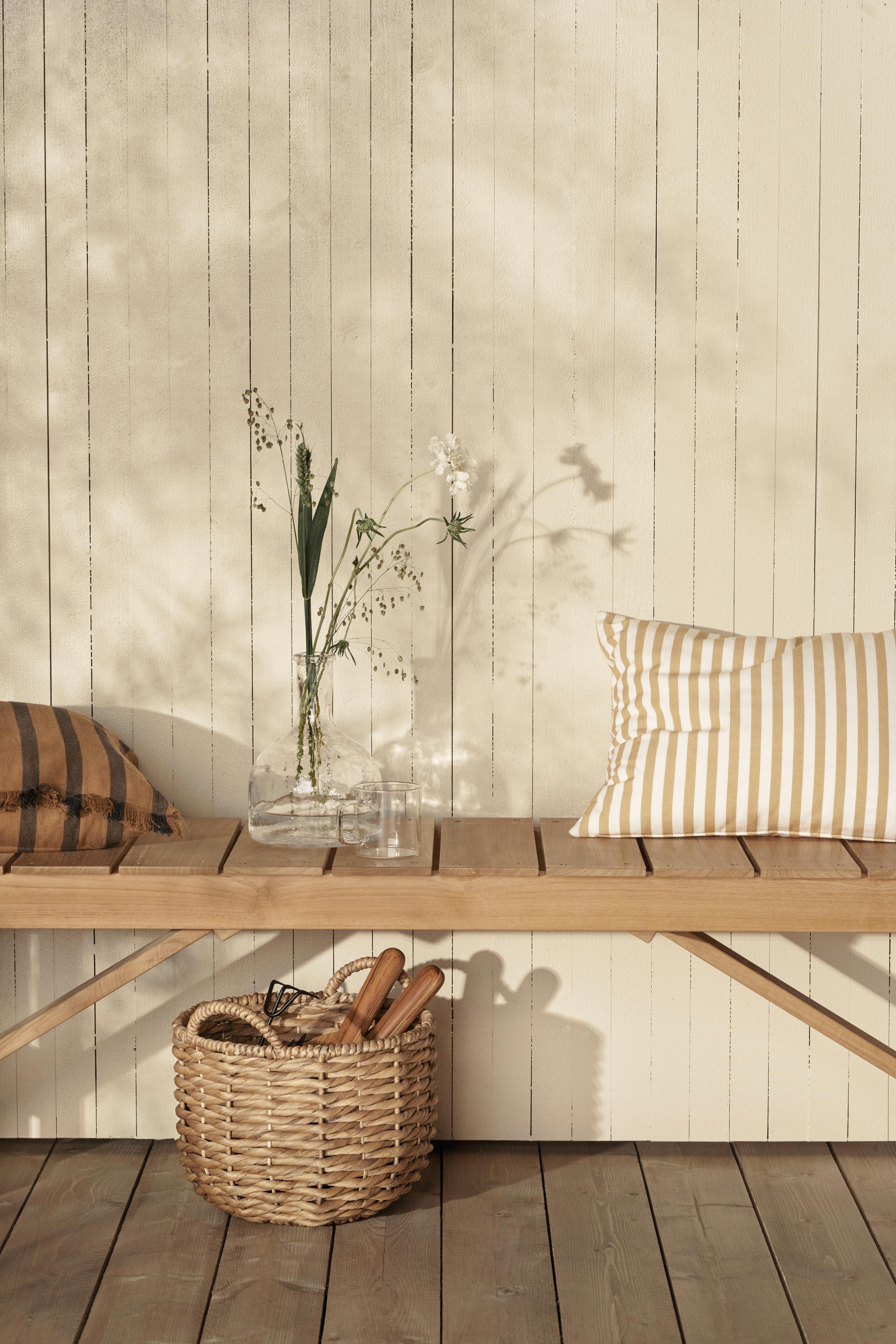 A wooden bench with striped cushions, a vase of flowers, and a woven basket of garden tools sits on a wooden deck against a paneled wall with soft foliage shadows.