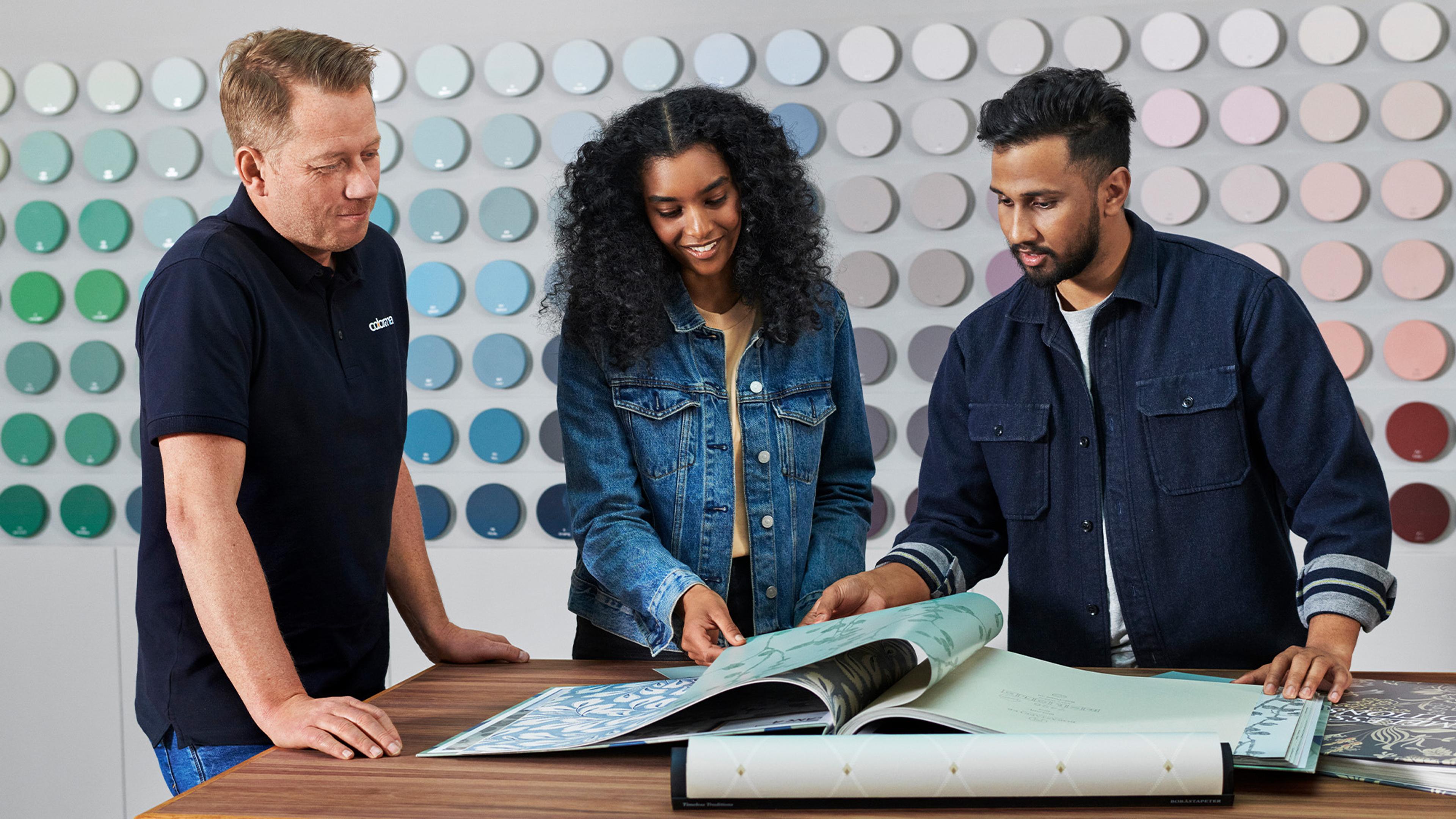 Three people looking at sample books at a table in a store with a wall of paint swatches.