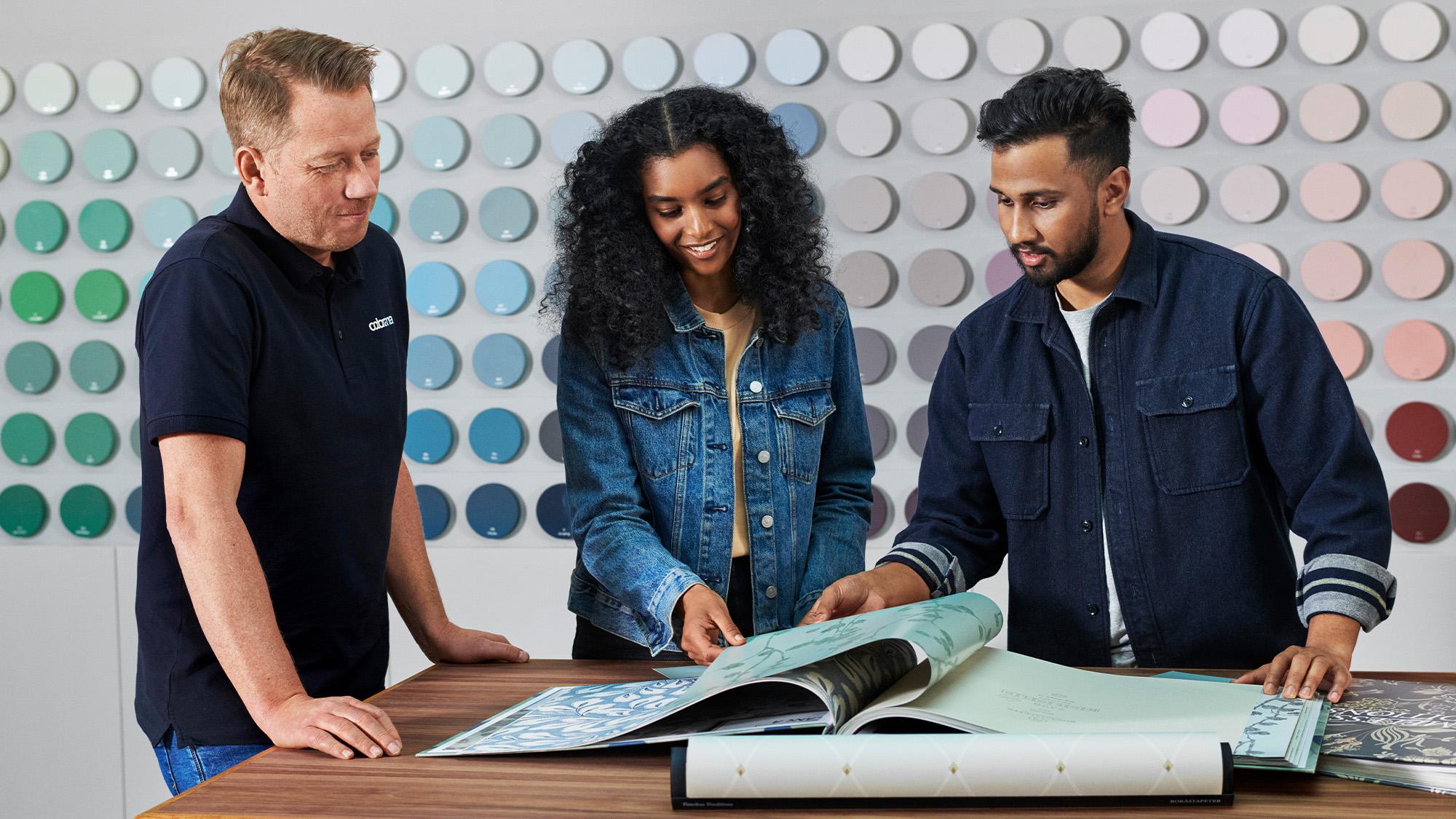 Three people examine design sample books in a store with a wall of colorful paint swatches.