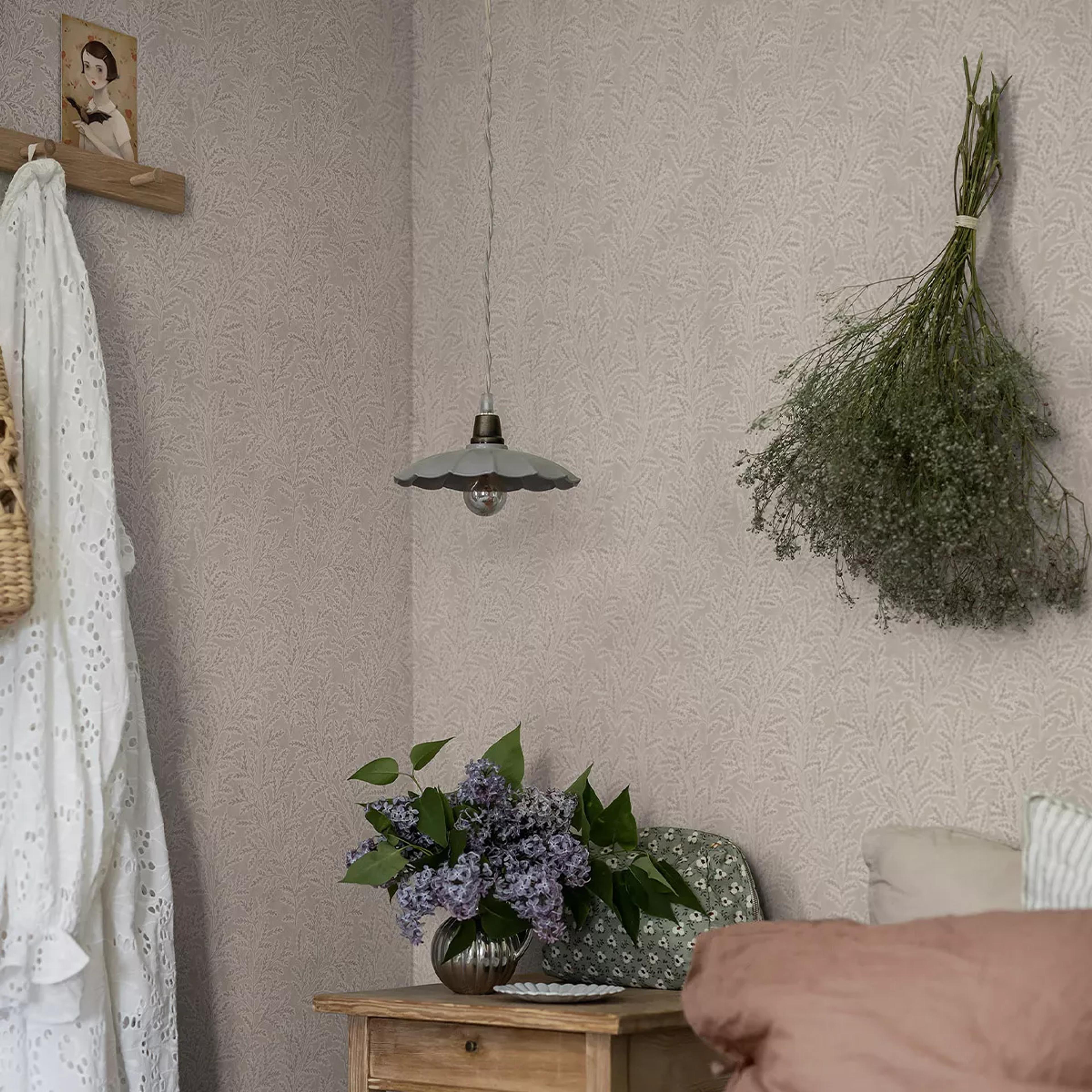 A cozy bedroom corner featuring light leaf-patterned wallpaper, a hanging lamp, dried flowers, a vase of lilacs on a wooden nightstand, and part of a bed.