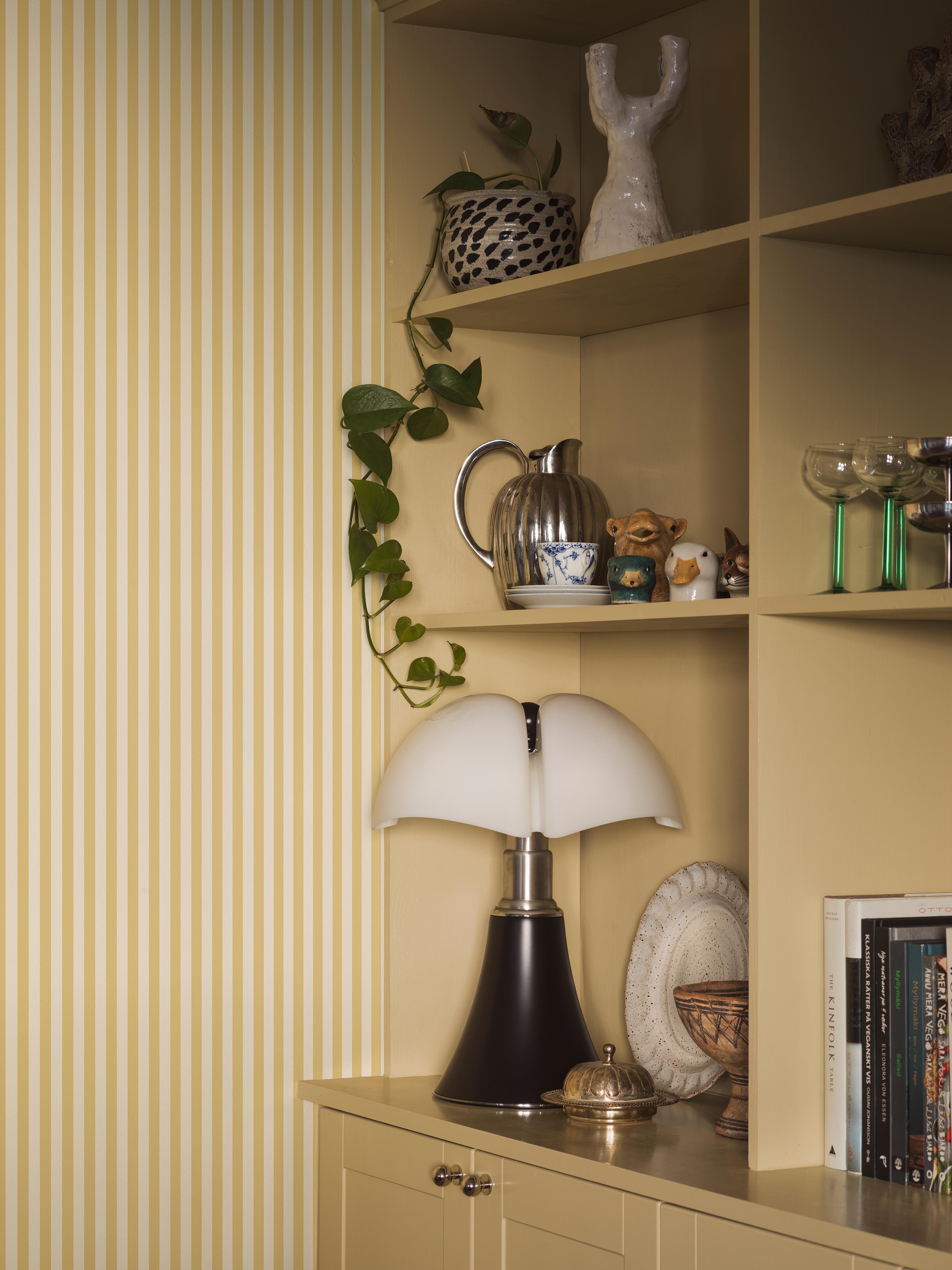 A yellow and white striped wall next to a yellow shelving unit filled with decorative items, including a vine plant, a silver pitcher, small sculptures, a mushroom-shaped lamp, and books.