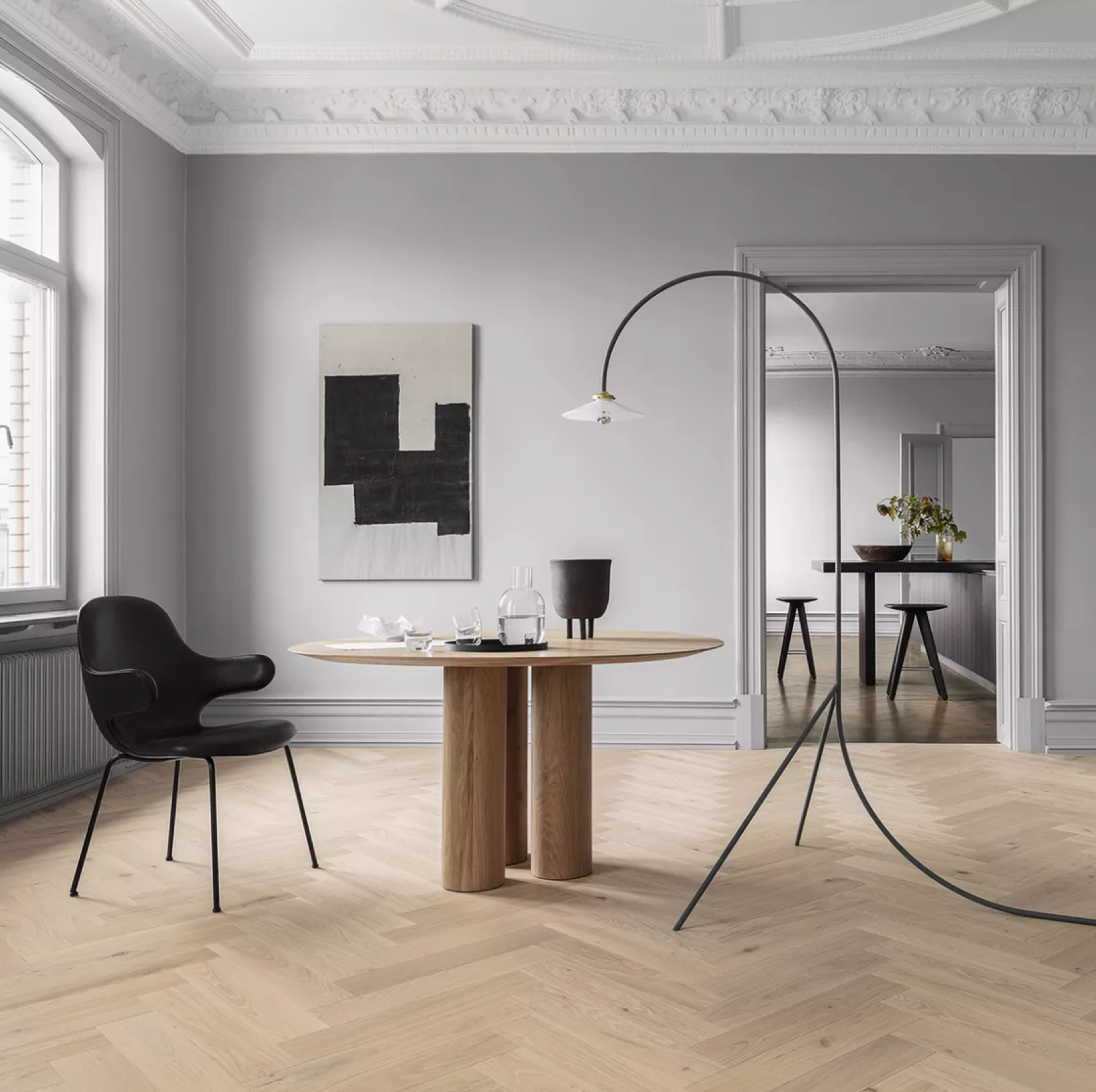 A modern dining room with light wood herringbone floors, a round wooden table, black chair, abstract art, and an arched floor lamp.