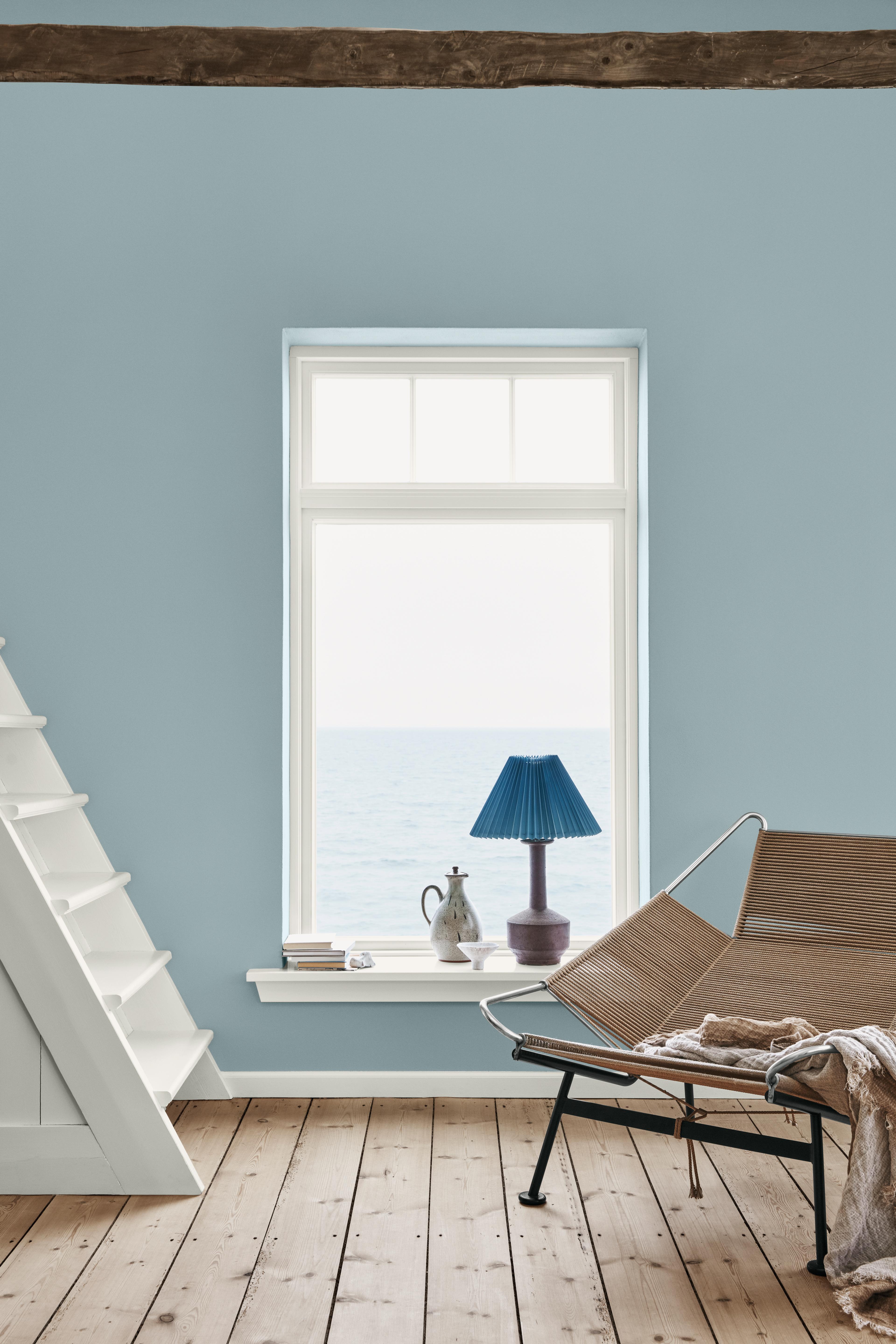 A light blue room with a large window overlooking the ocean, featuring a white staircase, a modern woven chair, and a blue lamp on the sill.