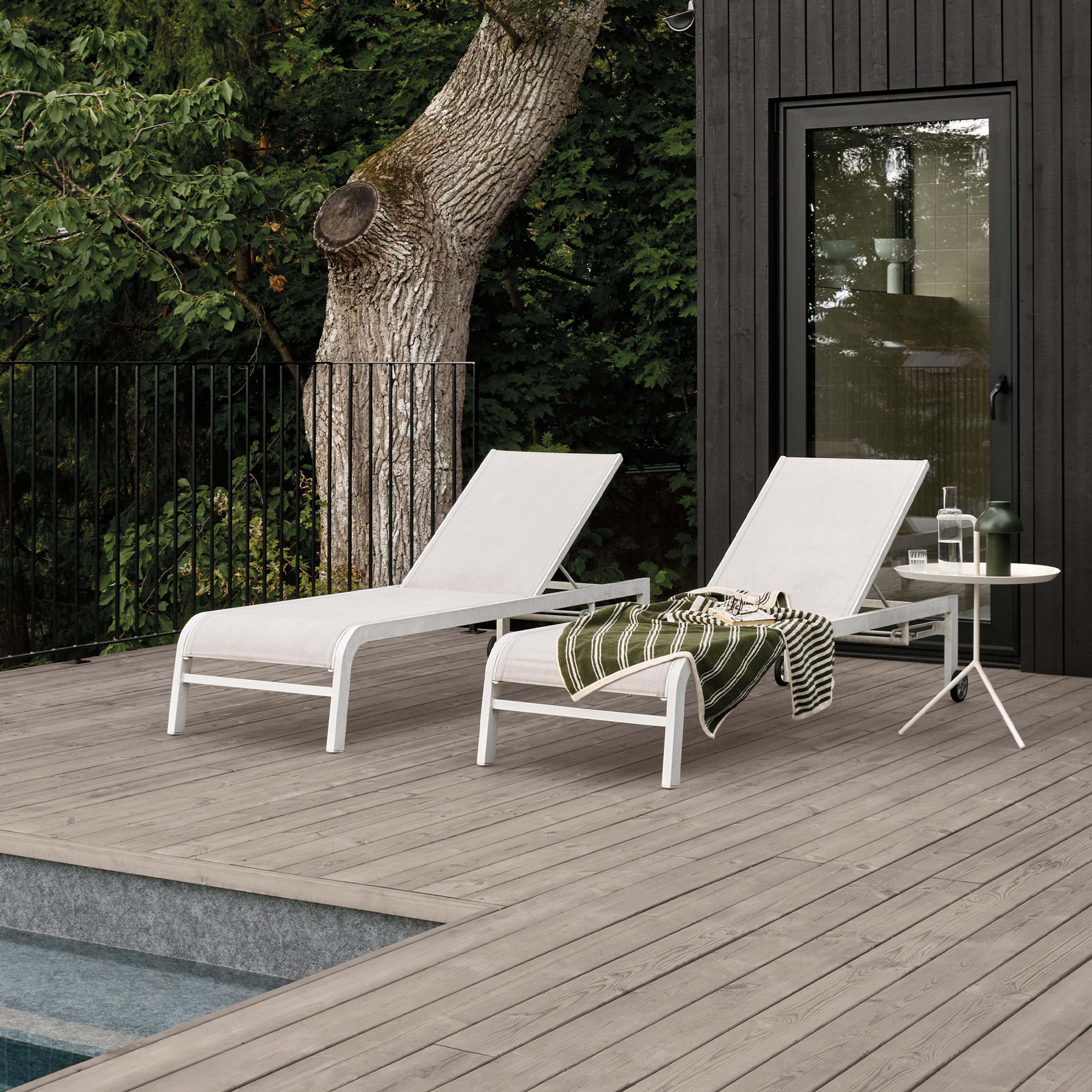Two white lounge chairs on a wooden deck next to a pool, with a modern black house and trees in the background.