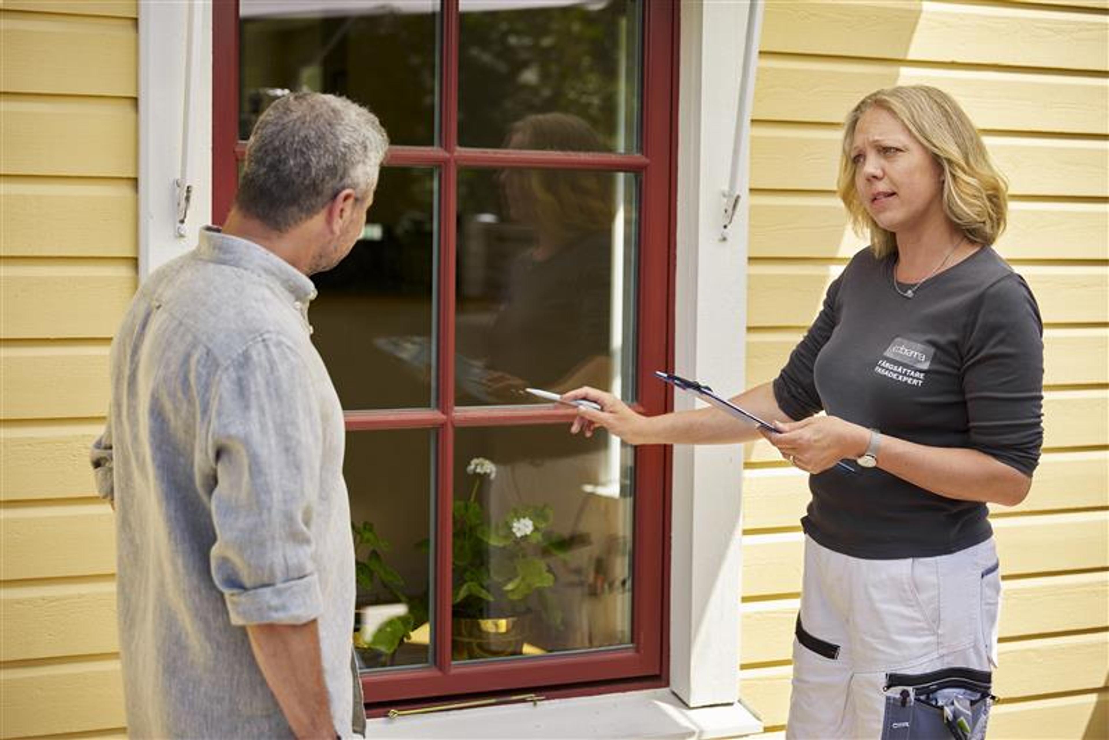A window expert points to a window on a yellow house while speaking to a man.