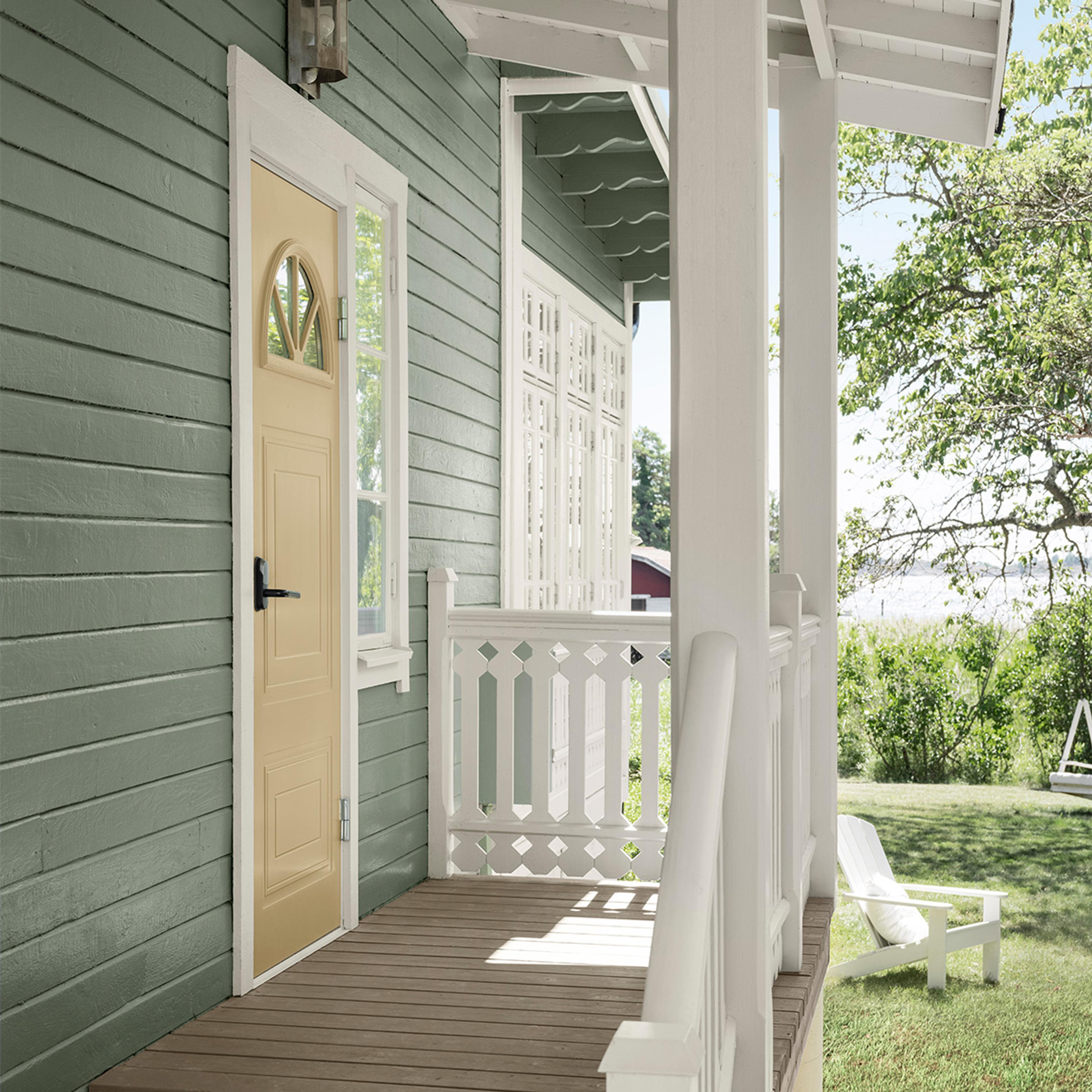 A light green house with a yellow door and white porch, overlooking a sunny yard with a lake.