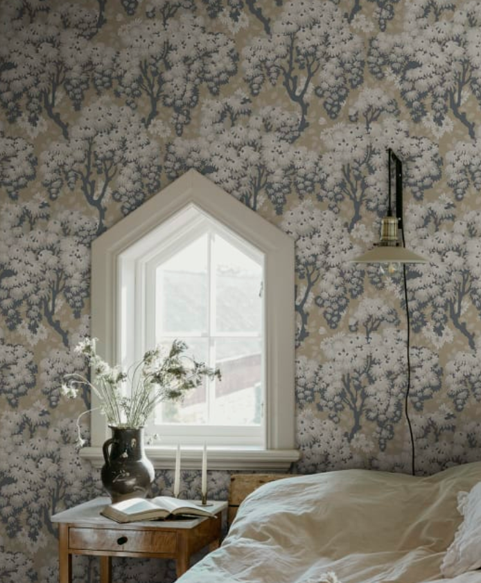 A bedroom featuring light brown wallpaper with a repeating pattern of gray trees, white foliage, and grape clusters. An arched window sits above a wooden nightstand holding flowers, an open book, and candles, with a bed visible in the foreground.