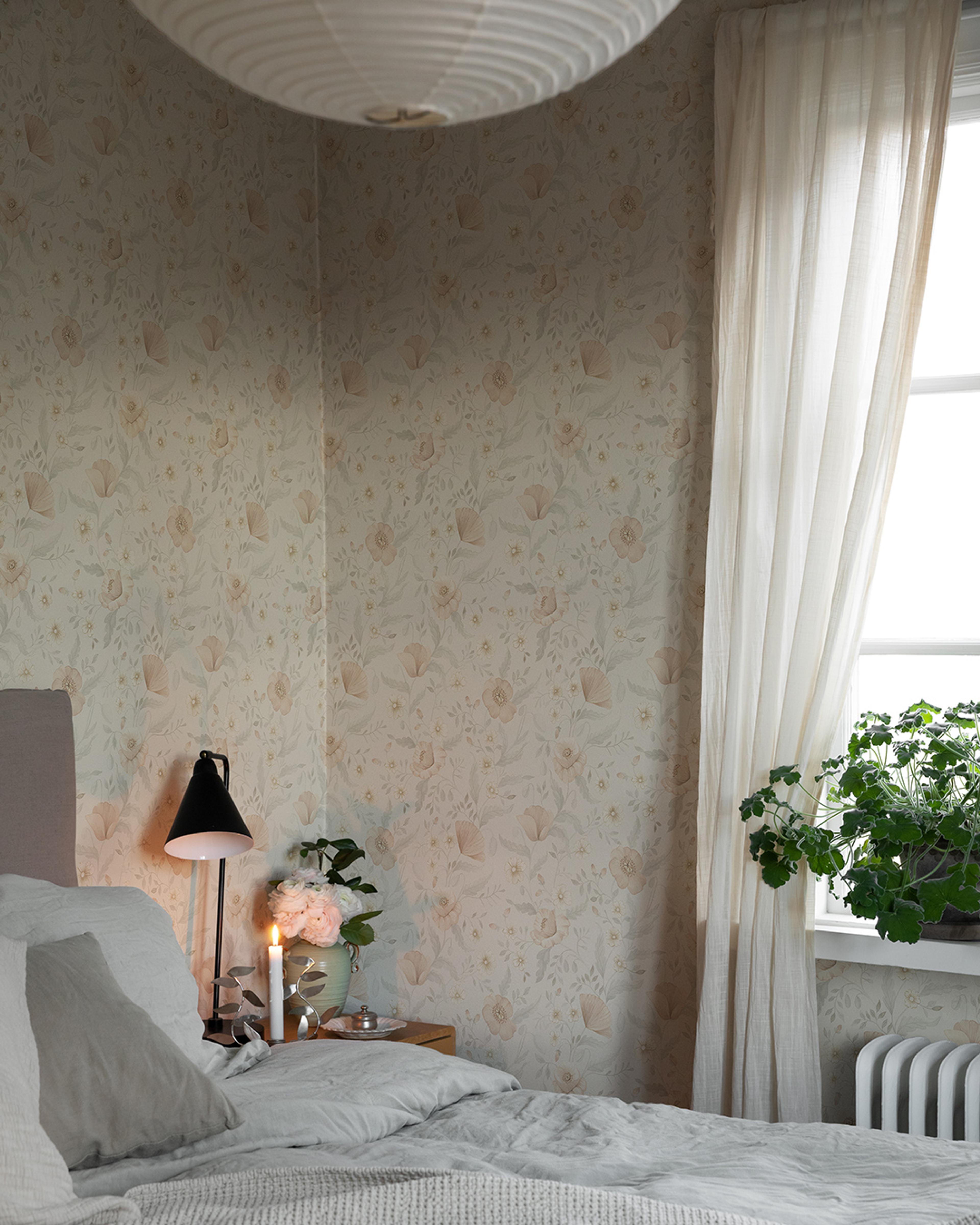 A serene bedroom with light floral wallpaper, a bed with white bedding, a lit lamp and candle on a bedside table with pink flowers, and a bright window with sheer curtains.