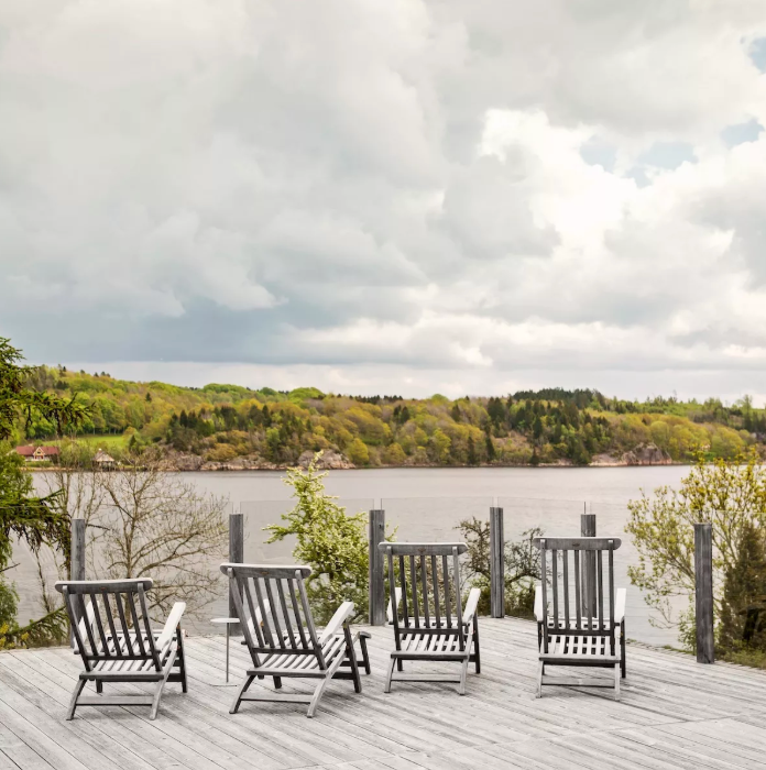 Lounge chairs on a wooden deck overlooking a lake with a forested shore under a cloudy sky.