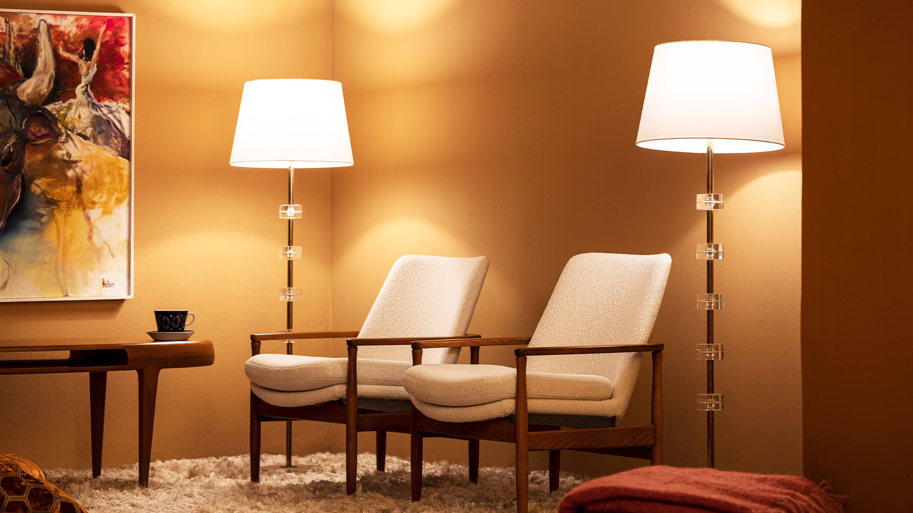 A warmly lit room with two cream armchairs, two tall floor lamps with crystal accents, an abstract painting, and a white shag rug.