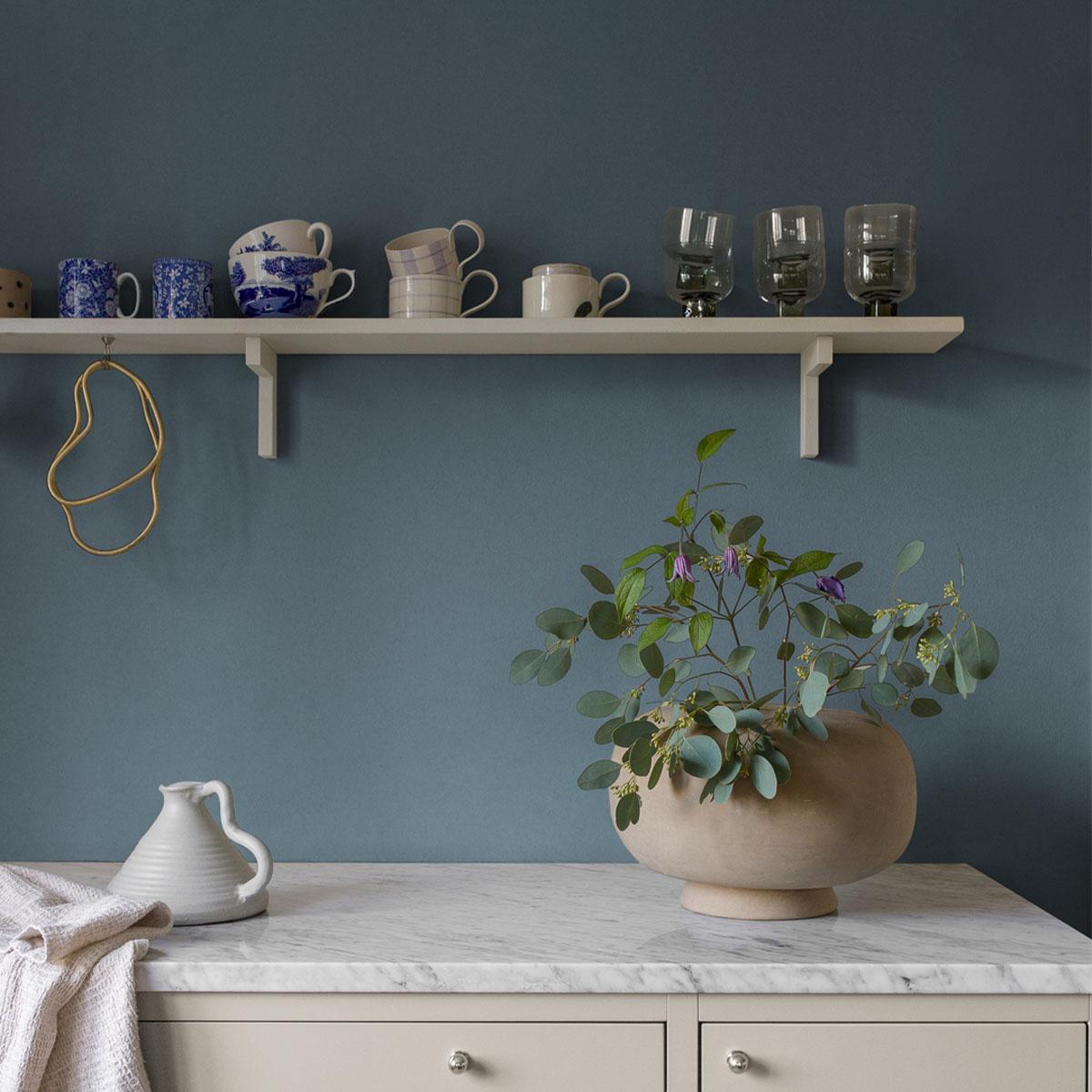 A white shelf on a blue wall holds various cups and glasses, while a marble counter below features a vase with eucalyptus and small purple flowers, a pitcher, and a towel.