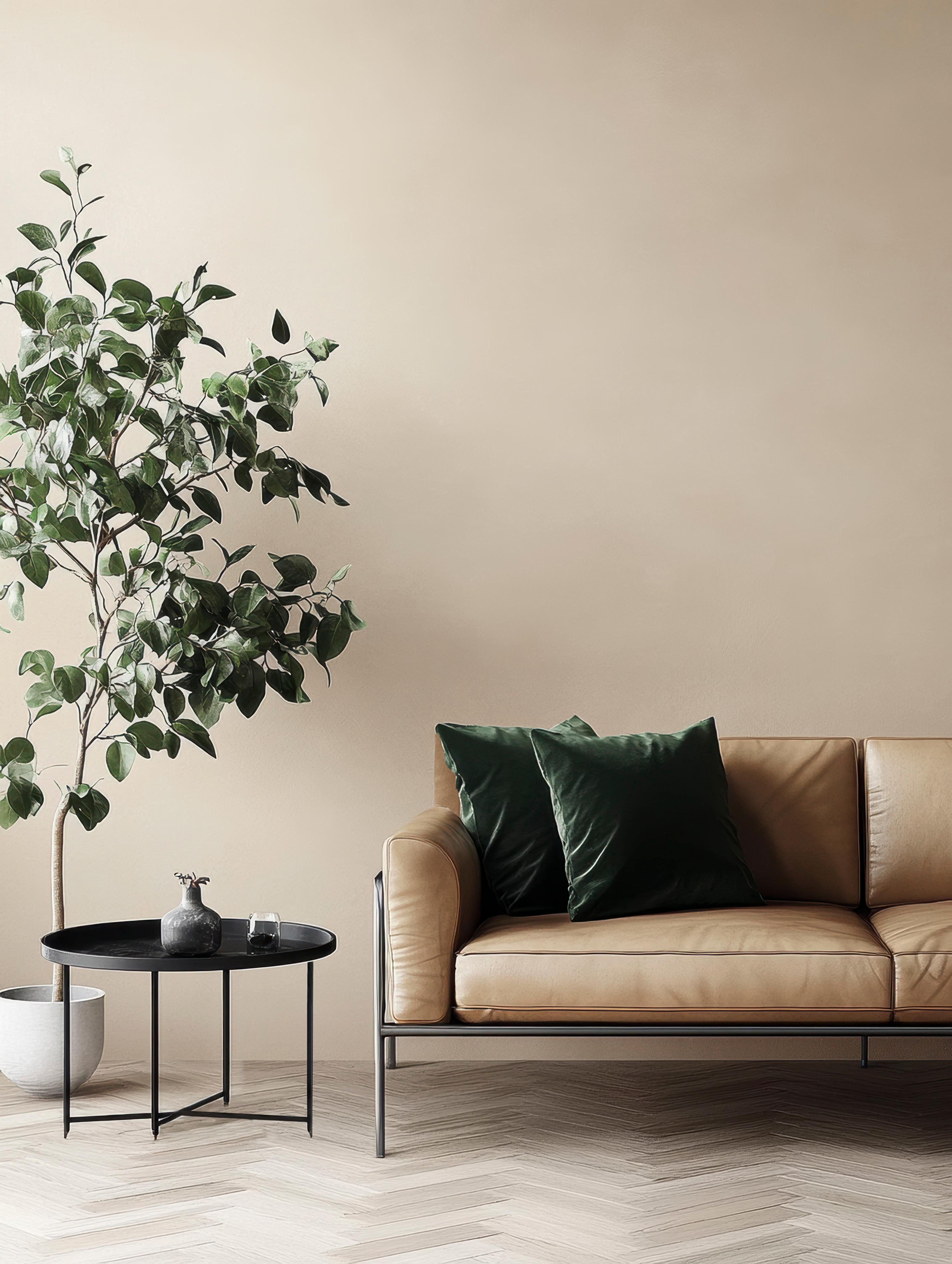 A modern living room with a tan leather sofa, two dark green velvet cushions, a black side table, and a large potted plant on a light herringbone floor.