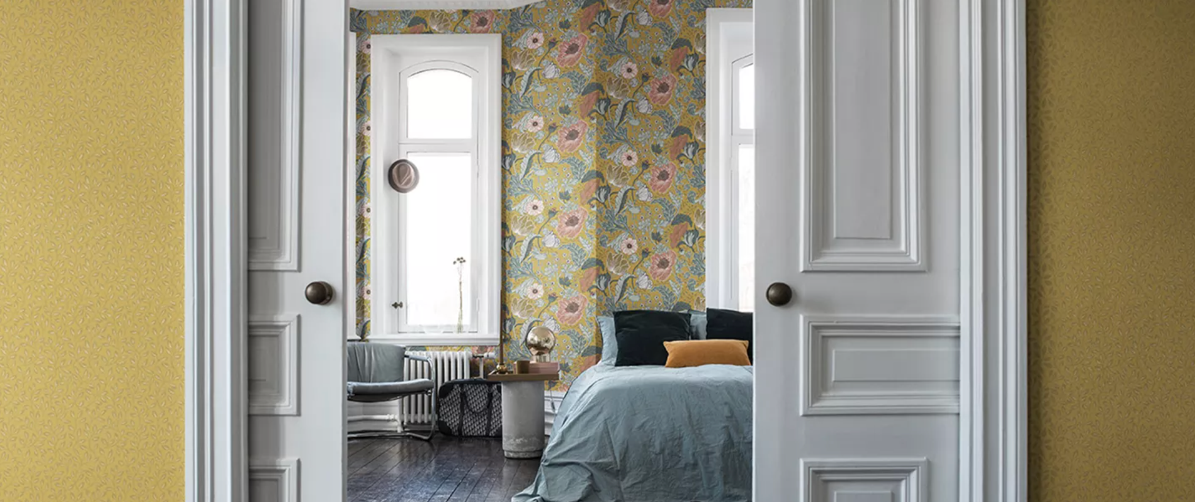 A bedroom with yellow-green floral wallpaper and a blue bed, seen through open white double doors from a yellow hallway.