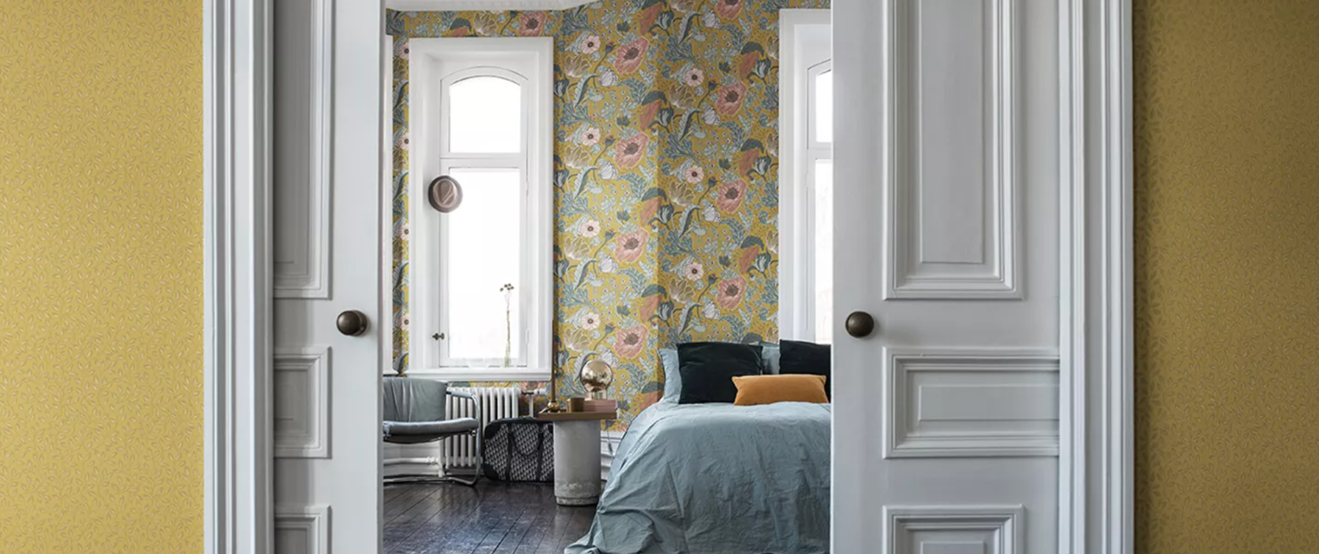 A bedroom with yellow-green floral wallpaper and a blue bed, seen through open white double doors from a yellow hallway.