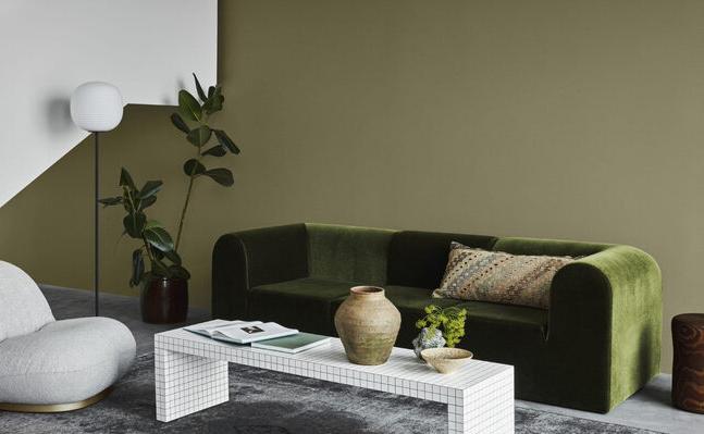 A modern living room with olive green and white walls, featuring a green velvet sofa, a grey sculptural armchair, and a grid-patterned coffee table on a grey rug.
