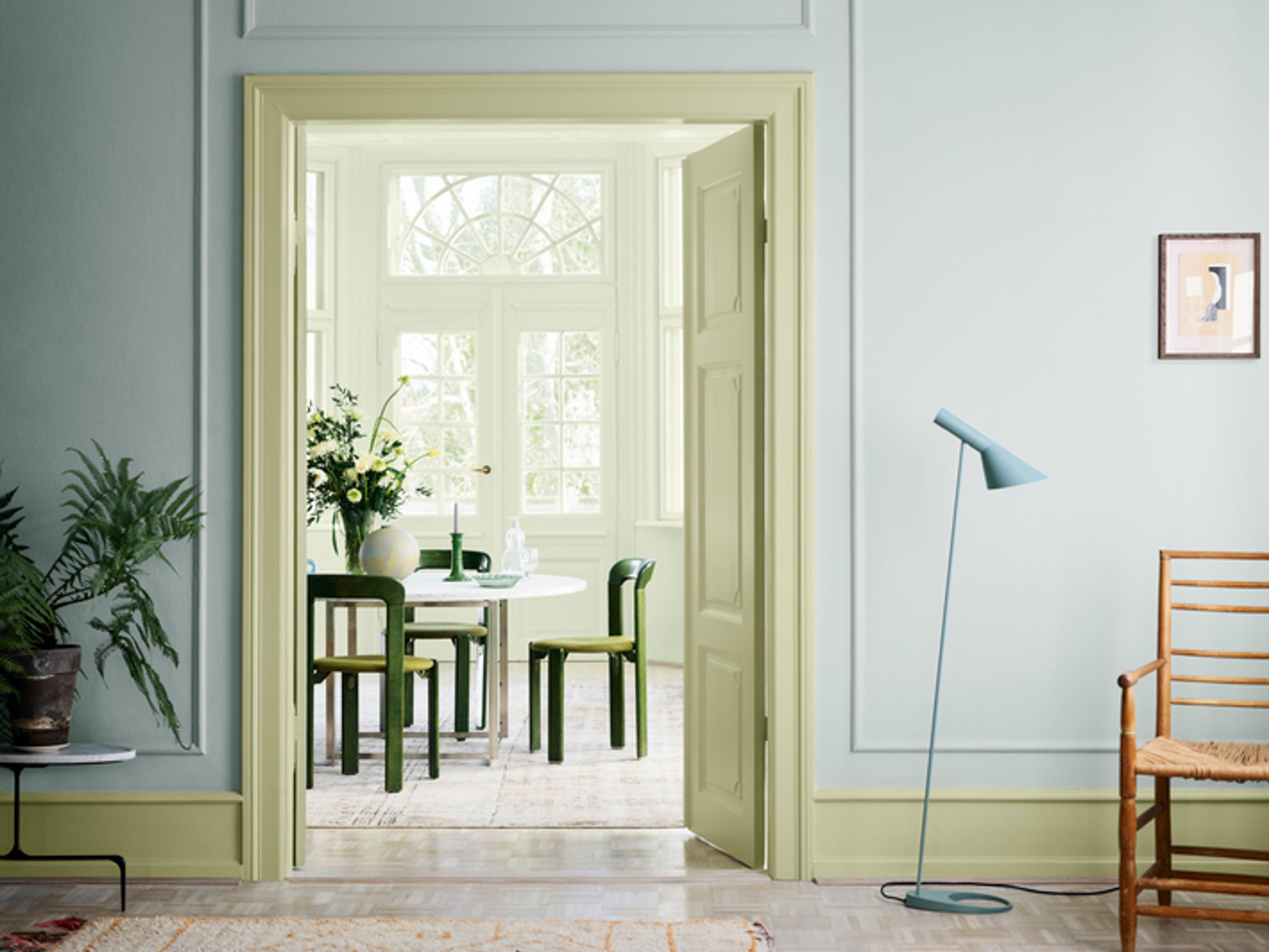 A light blue room with light green trim, looking through a doorway into a light green dining room with a white table and dark green chairs.