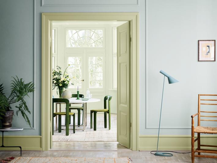 A light blue room with light green trim, looking through a doorway into a light green dining room with a white table and dark green chairs.