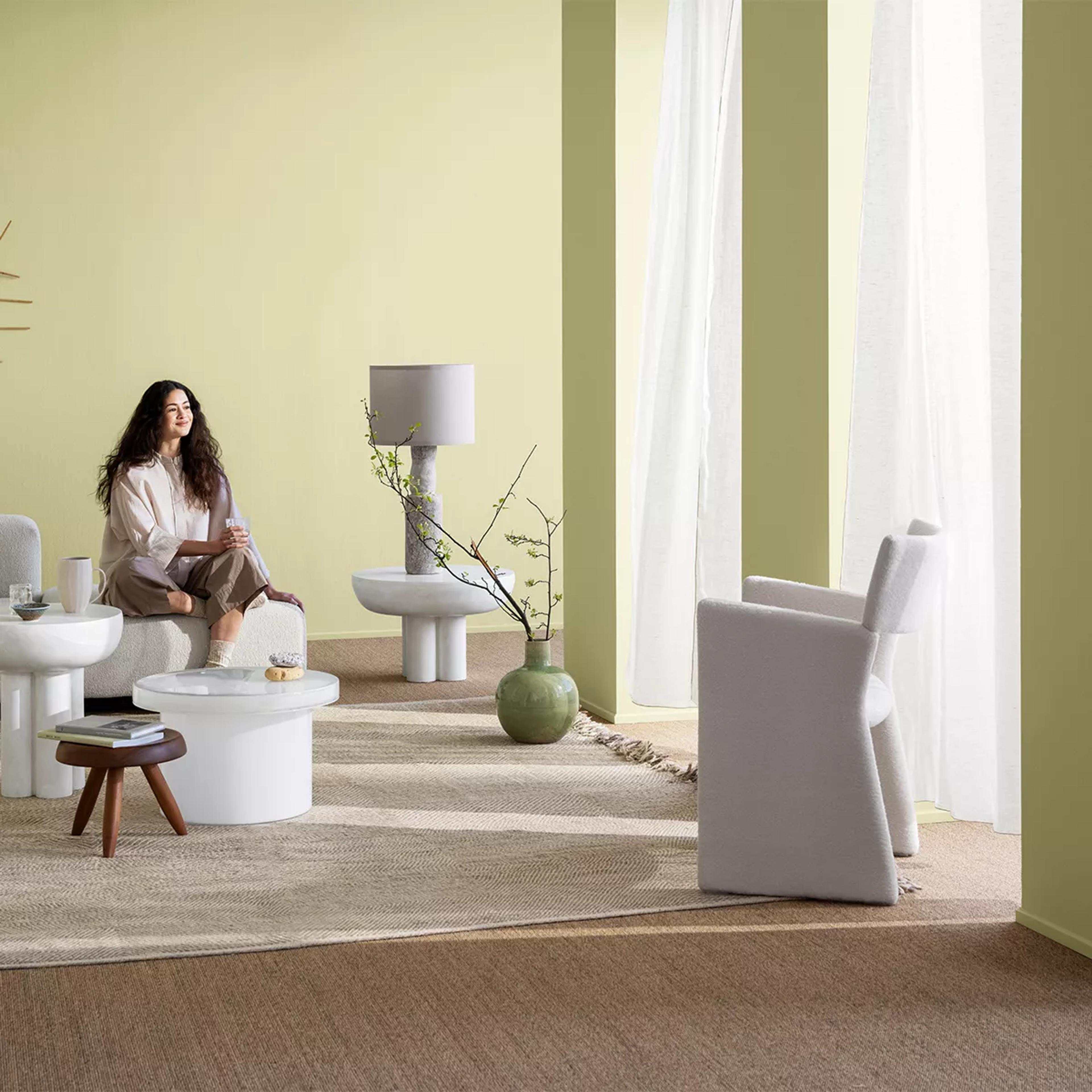 A woman sits on a textured white bench in a living room with pale green walls, modern tables, and a grey armchair.
