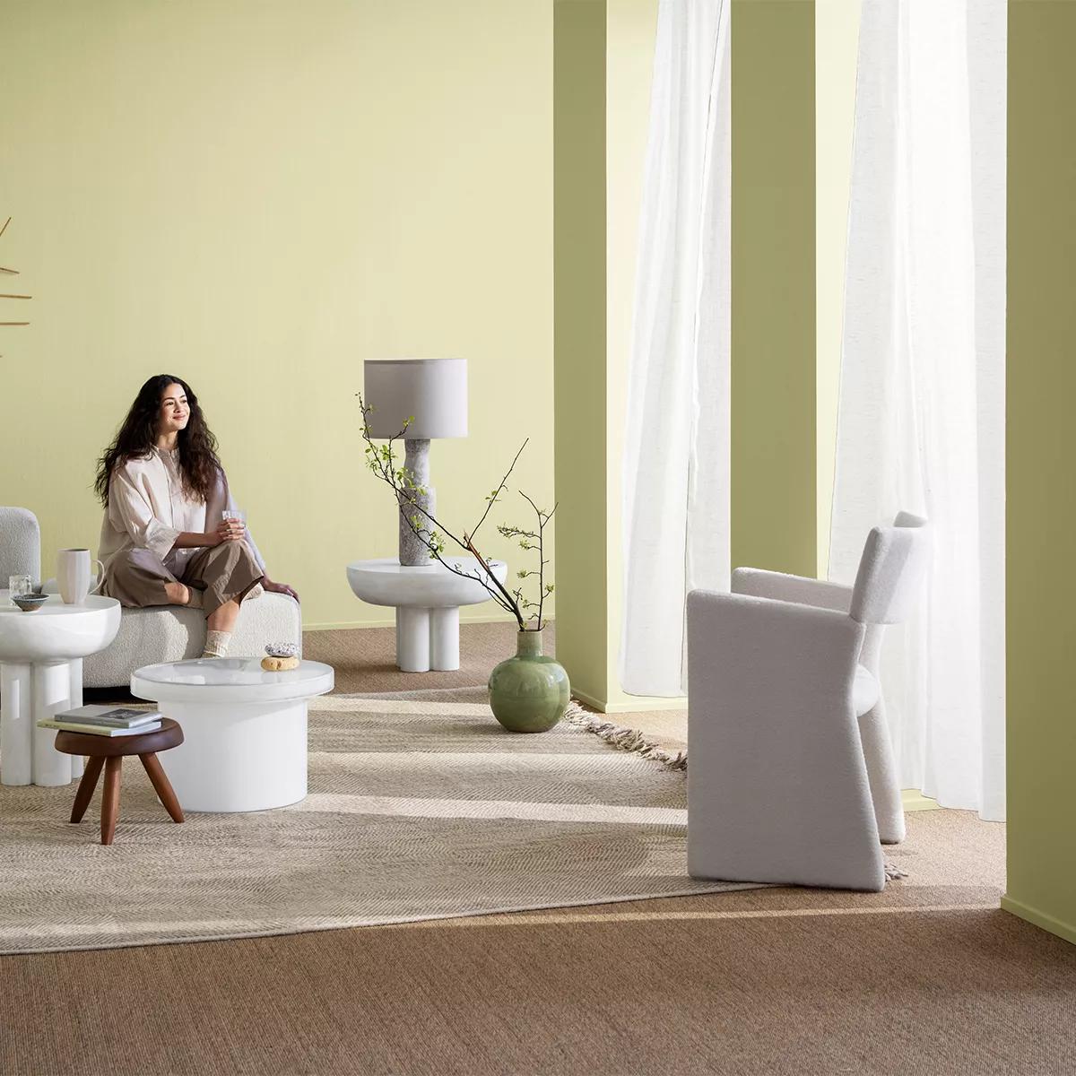 A woman sits on a textured white bench in a living room with pale green walls, modern tables, and a grey armchair.
