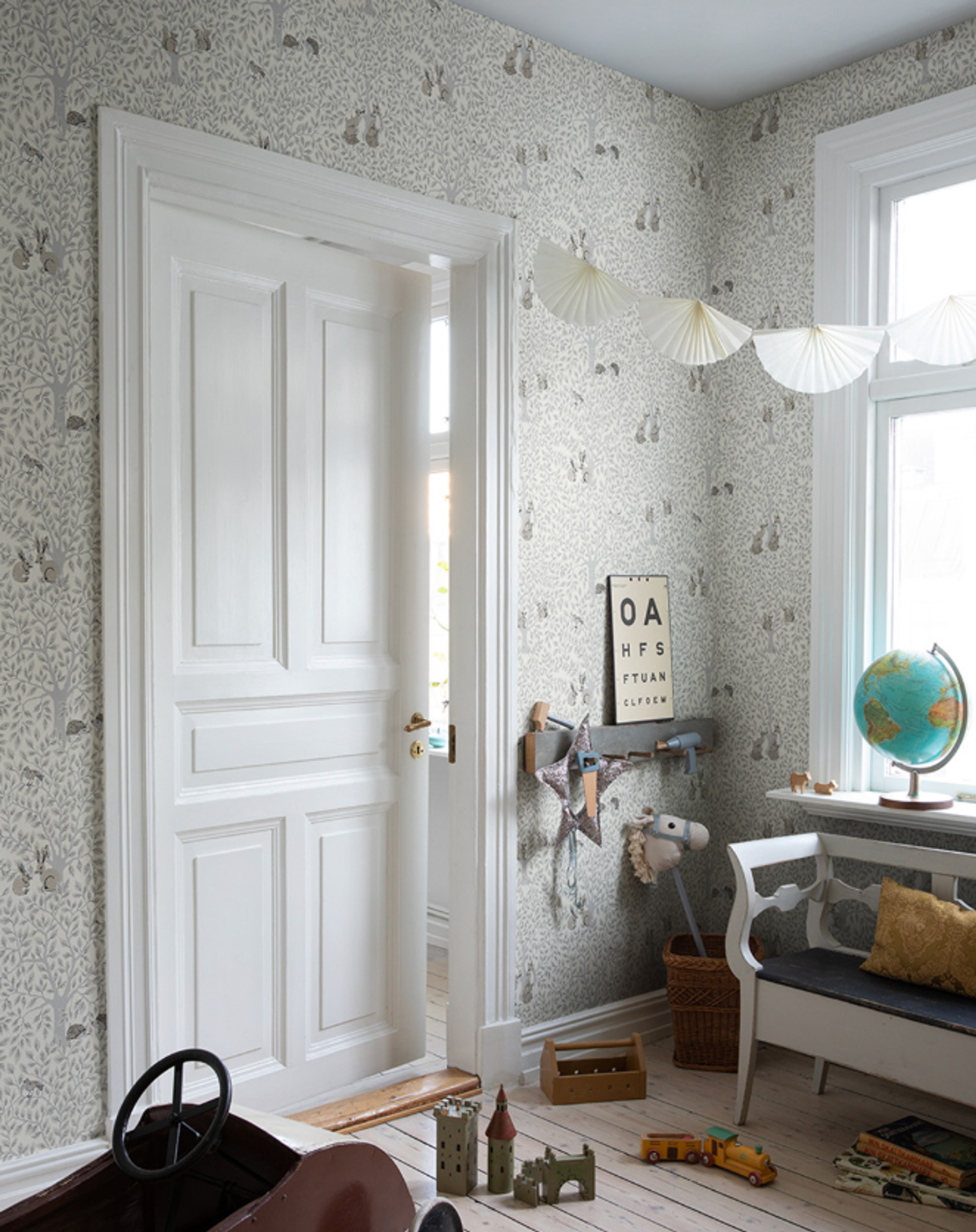 A light-filled child's room with forest animal wallpaper, a white door slightly ajar, a bench, a globe, and various toys on a pale wooden floor.
